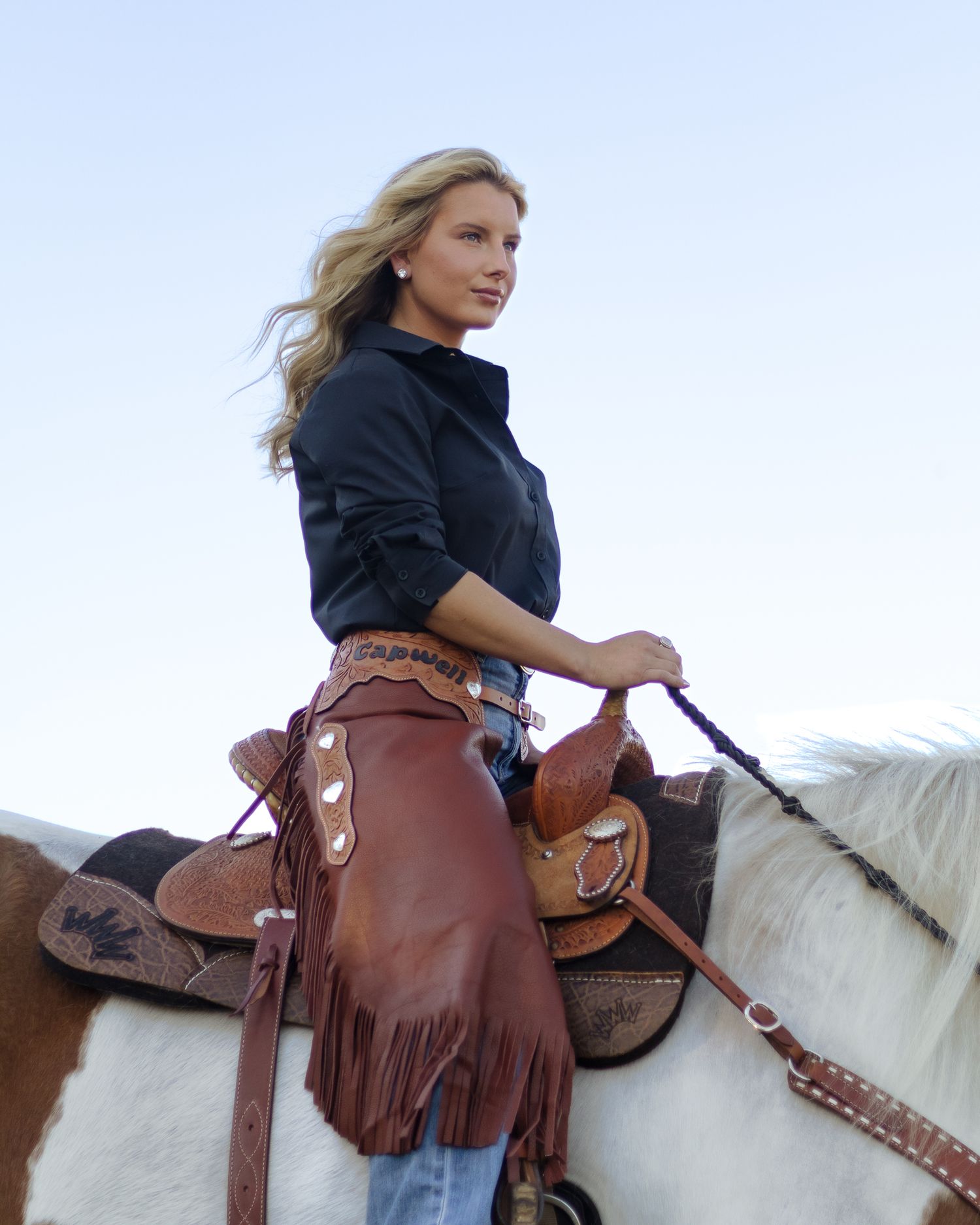 Woman with long blonde hair rides a white and brown horse, wearing a dark button-up shirt, fringed brown leather chaps and a western saddle.