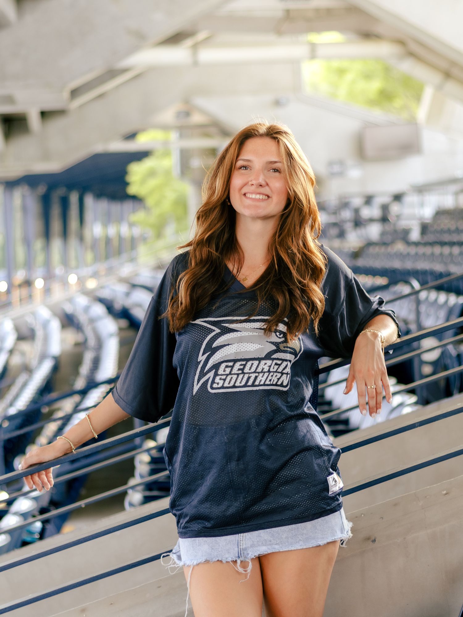 Smiling young woman in a navy Georgia Southern jersey and denim shorts poses on the stadium steps with empty bleachers behind her.