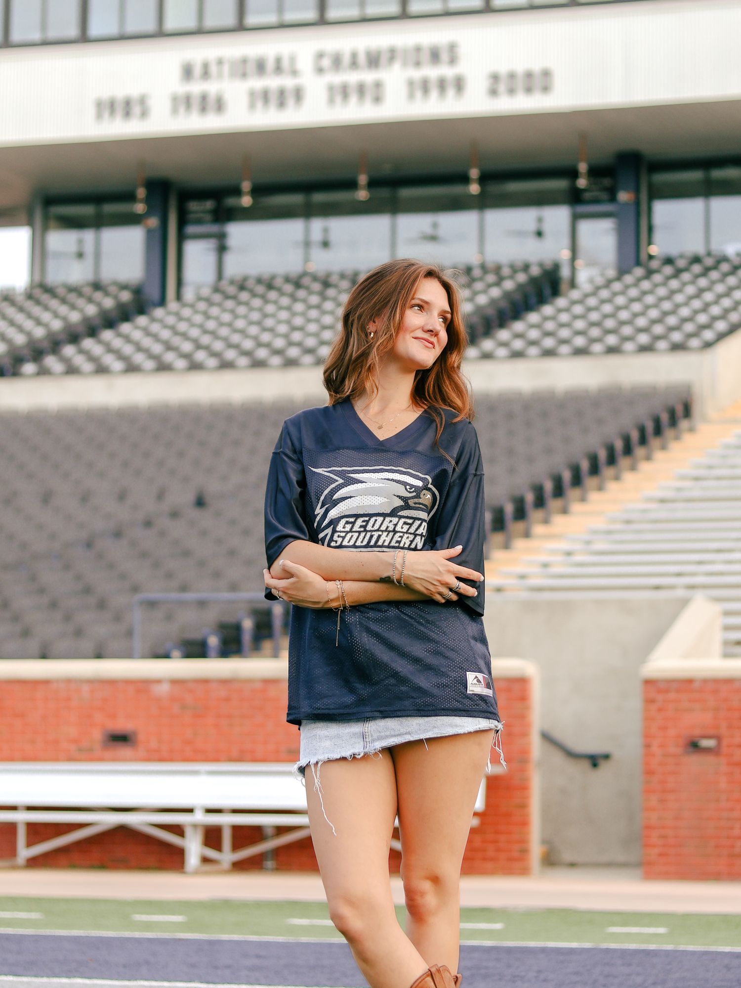 Woman in a Georgia Southern eagle jersey and denim shorts poses with crossed arms in a stadium with national champions banner behind her.