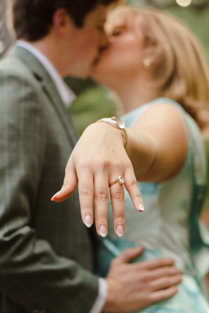 Couple kissing in the background while the woman extends her hand in focus to show off a sparkling diamond engagement ring.