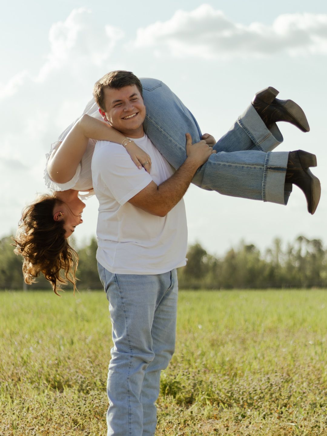 Smiling young man in a white t-shirt and jeans lifts a laughing woman in cowboy boots upside down over his shoulder in a sunny grassy field.