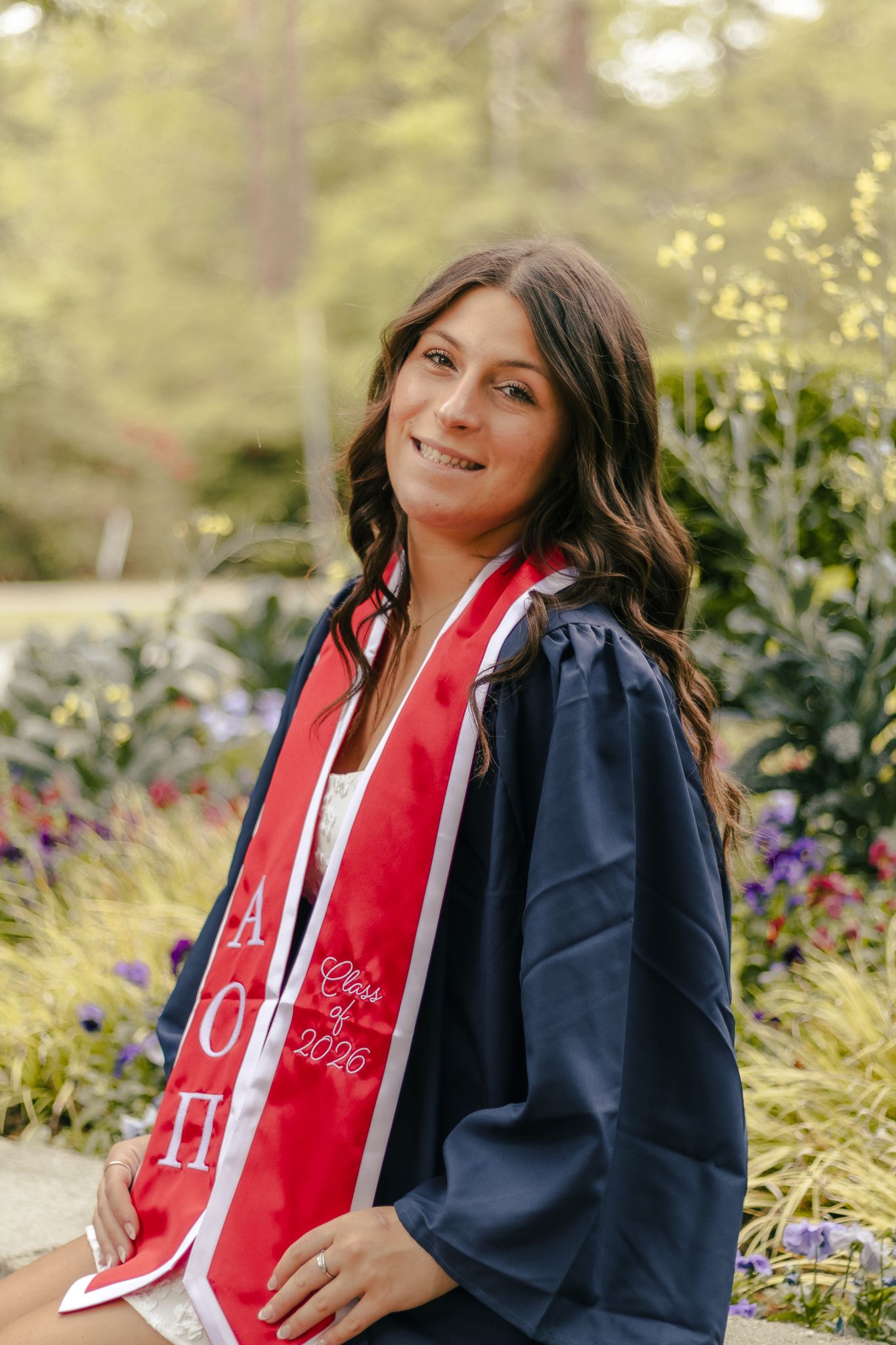 Smiling graduate in a navy gown and red Class of 2026 stole with Greek letters sits among colorful flowers in a garden.