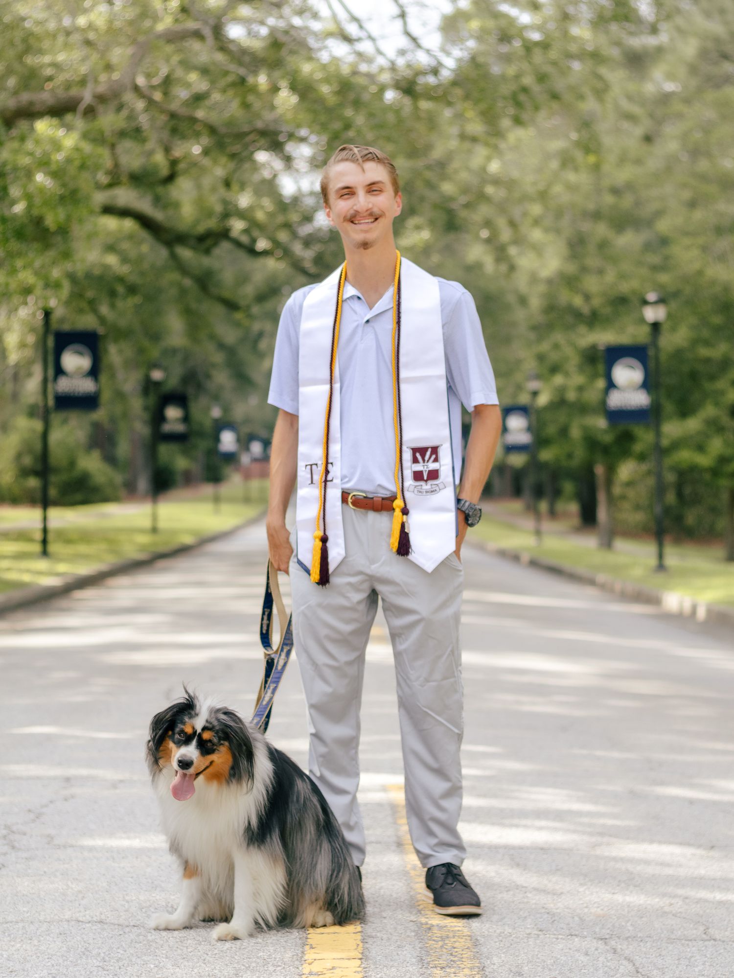 Smiling graduate in a white honor stole stands on a tree-lined campus road beside his panting Australian Shepherd on a leash.