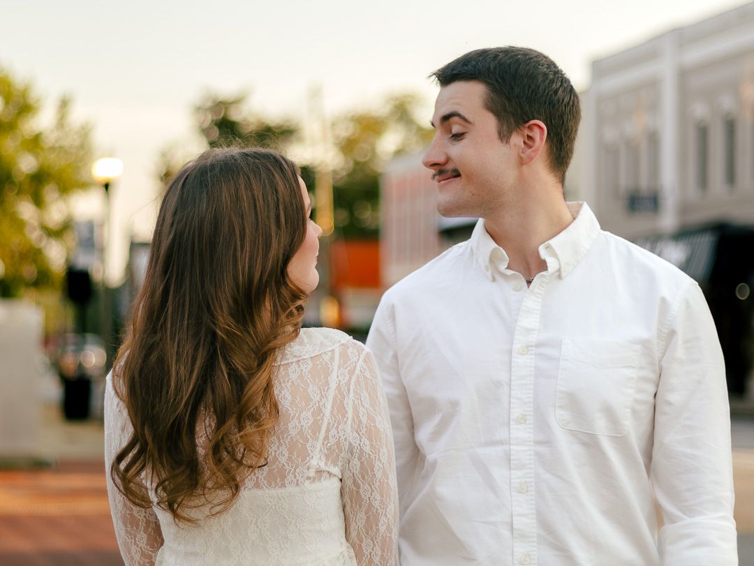 Smiling man in a white button-up shirt standing close to a woman with long brown hair in a lace dress on a city street.