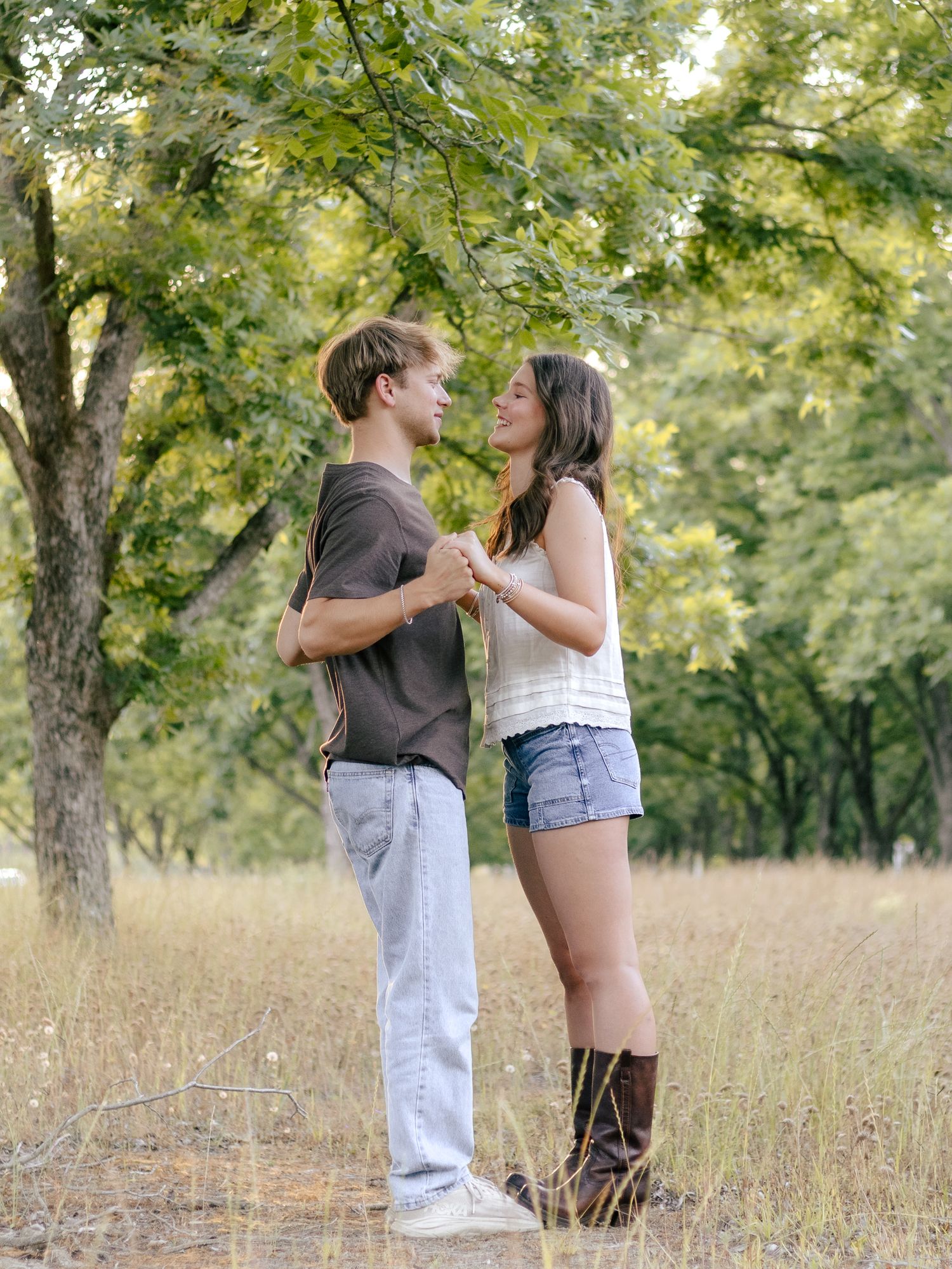 Young couple holding hands and smiling at each other while standing in a sunny grassy field beneath leafy green trees.