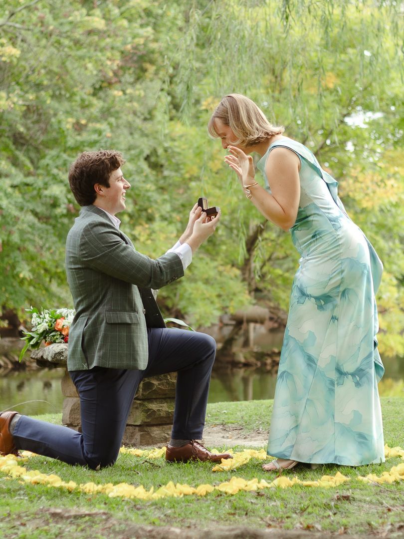 A woman in a flowing teal dress covers her mouth in joyful surprise as a kneeling man presents an engagement ring in a leafy park.