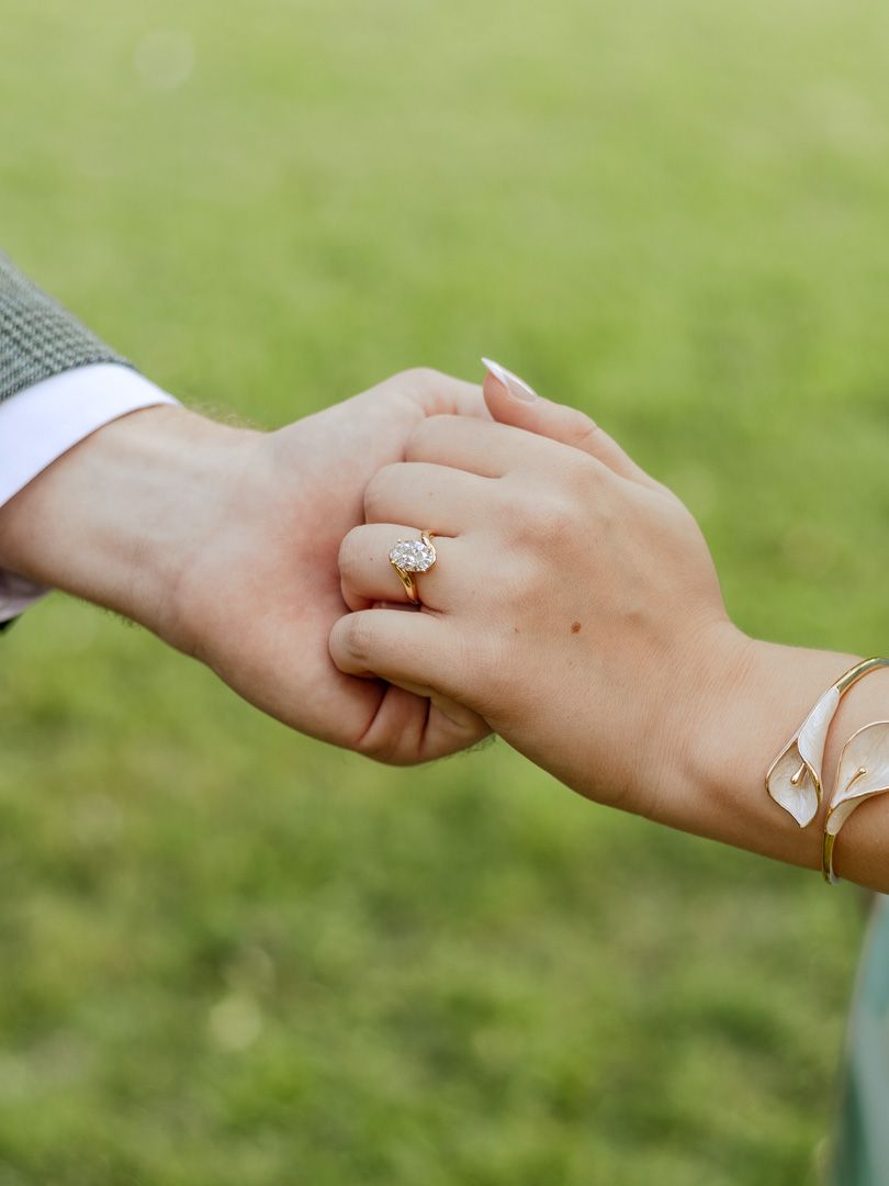 Close-up of a couple holding hands on a green lawn, showing a sparkling diamond engagement ring and a gold bracelet.