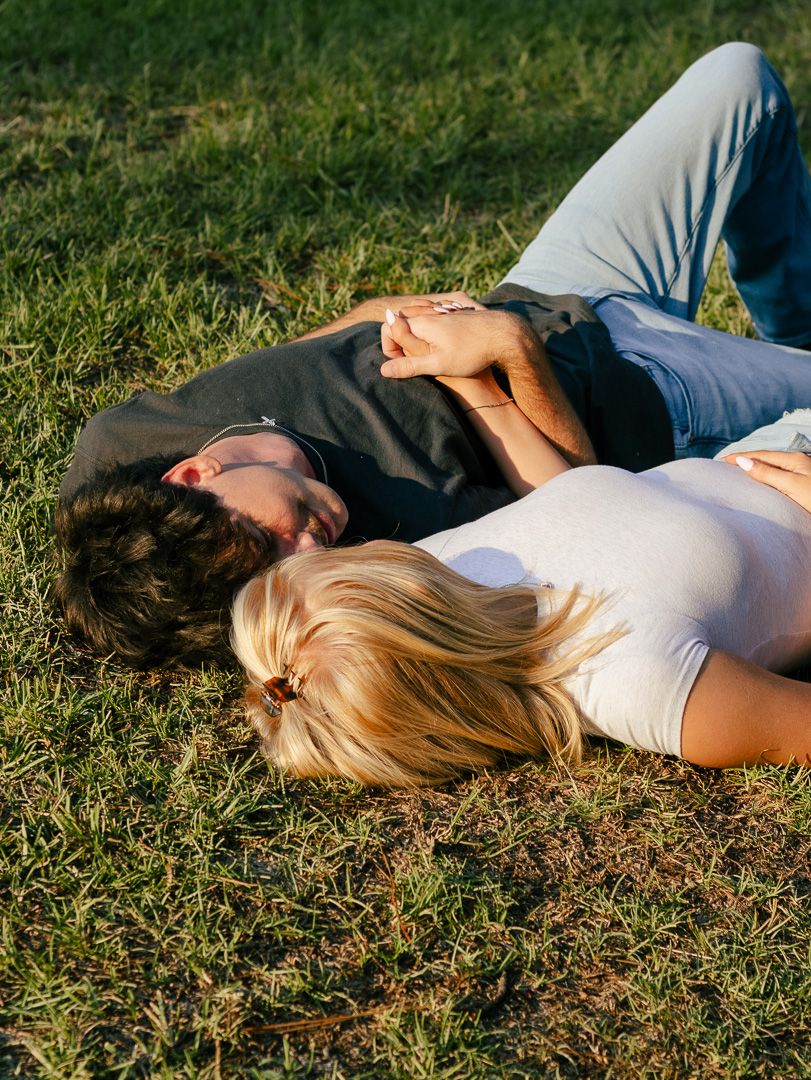 A young couple lies close together on green grass, embracing and relaxing outdoors on a sunny day.
