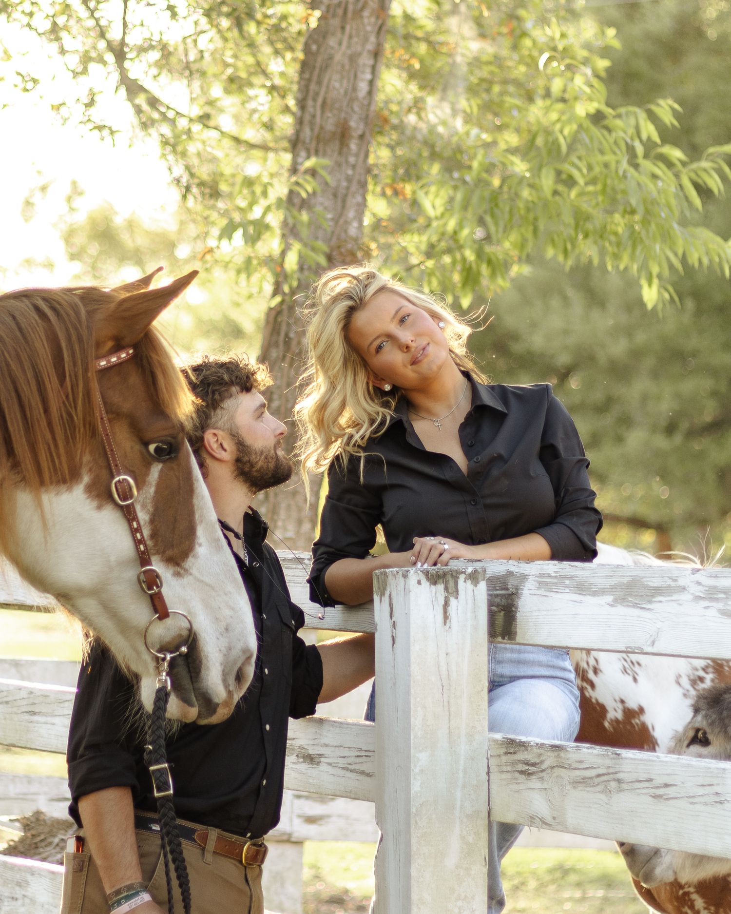 A smiling blonde woman and a bearded man in dark shirts lean on a white wooden fence beside a brown and white horse outdoors.