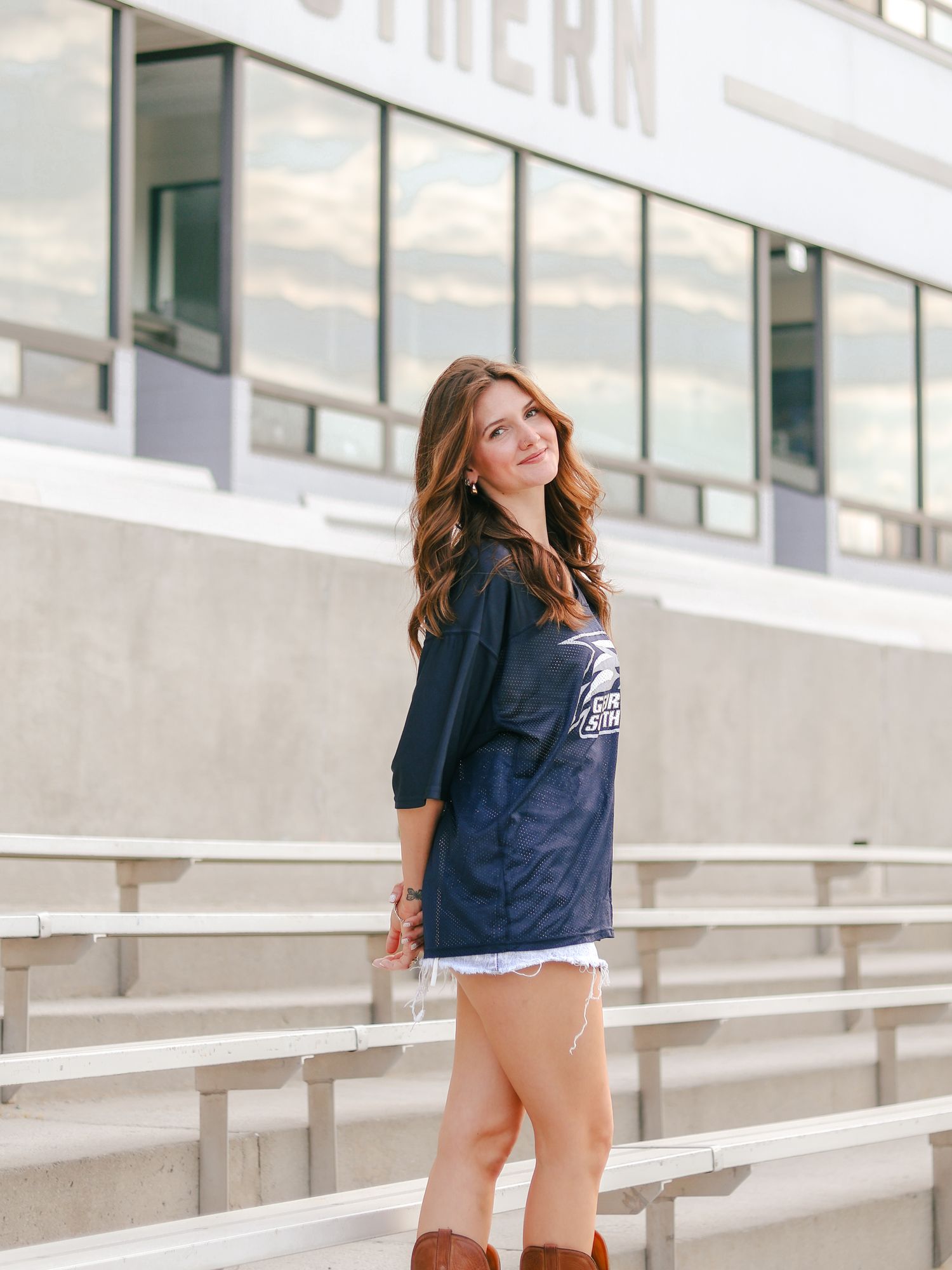 Woman with long wavy hair smiles over her shoulder in a navy jersey, denim shorts and brown boots on empty aluminum stadium bleachers.