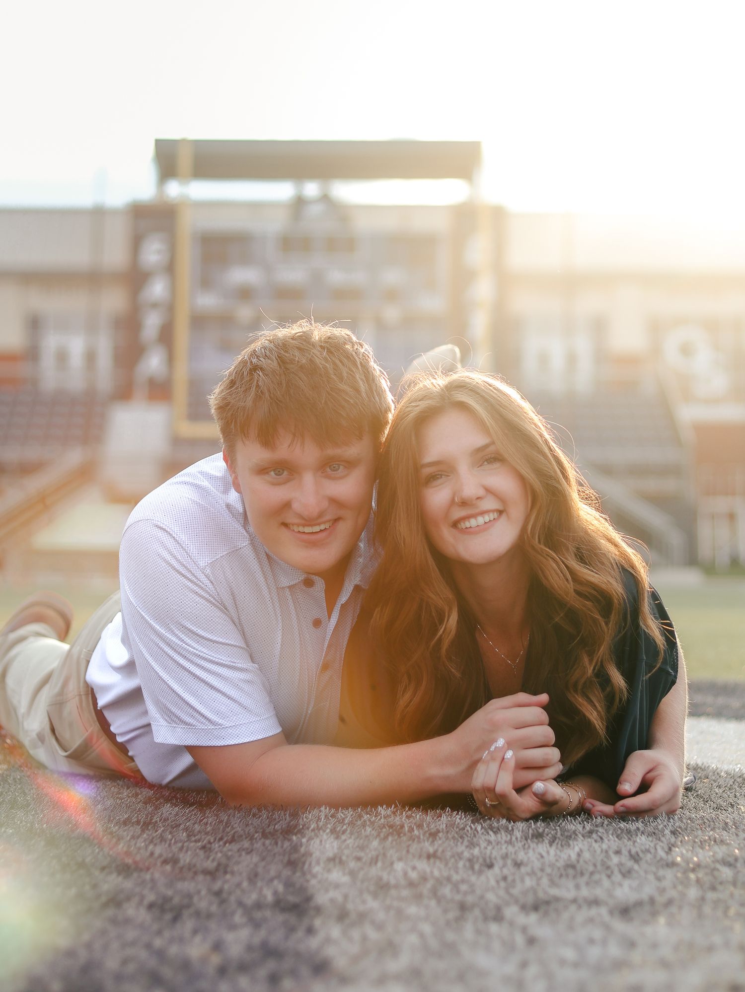 Smiling couple lying side by side on a grassy field, holding hands during a golden hour engagement photo session with a stadium behind them.