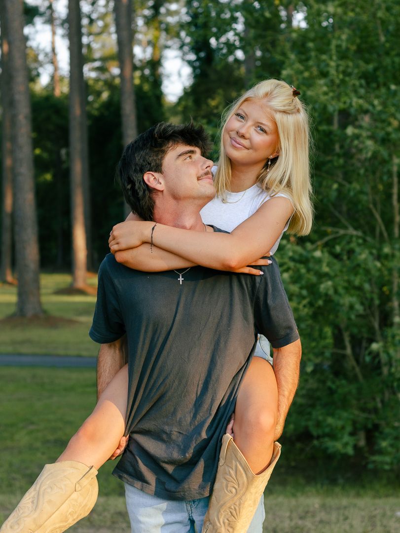 A smiling blonde woman in tan cowboy boots rides piggyback on a young man in a gray t-shirt on a grassy lawn surrounded by trees.