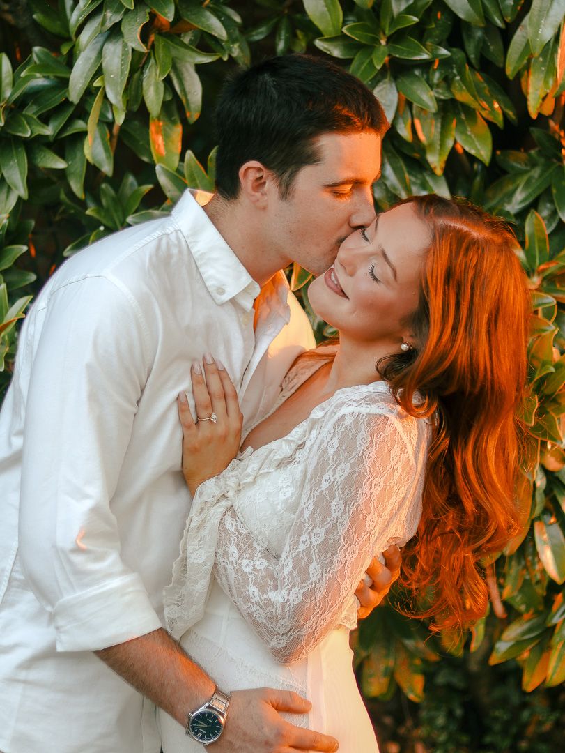 A man in a white shirt kisses the cheek of a smiling red-haired woman in a white lace dress, surrounded by green leaves.