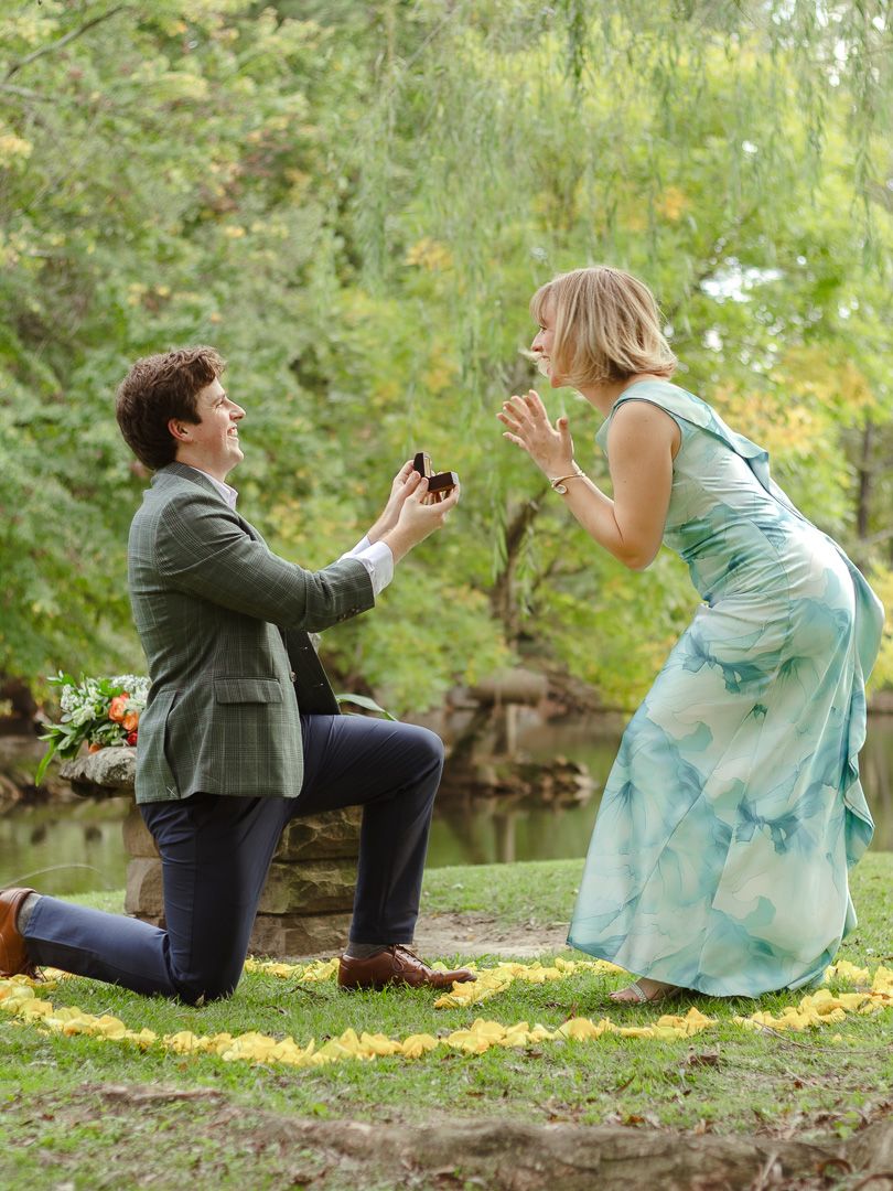 A man in a green blazer kneels on one knee holding a ring box as a smiling woman in a teal dress reacts during a park marriage proposal.