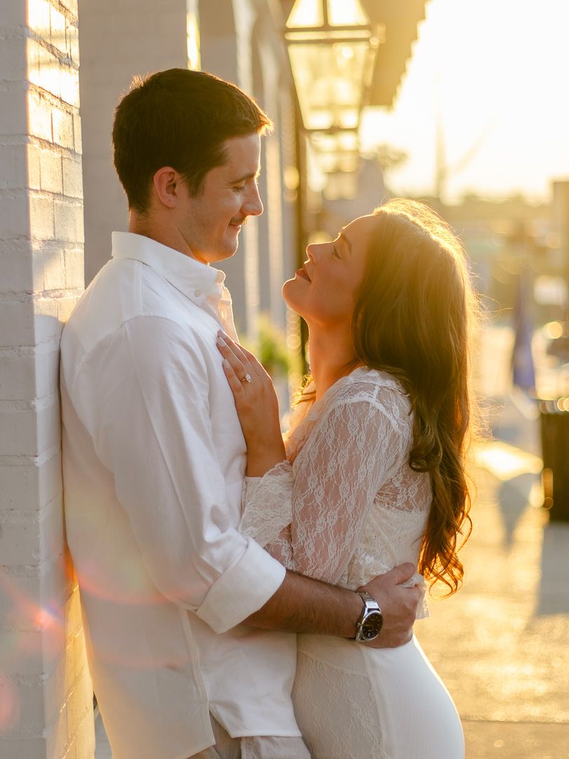 Couple embracing in golden sunset light on a city street, the woman gazing up at the man leaning against a white brick wall.