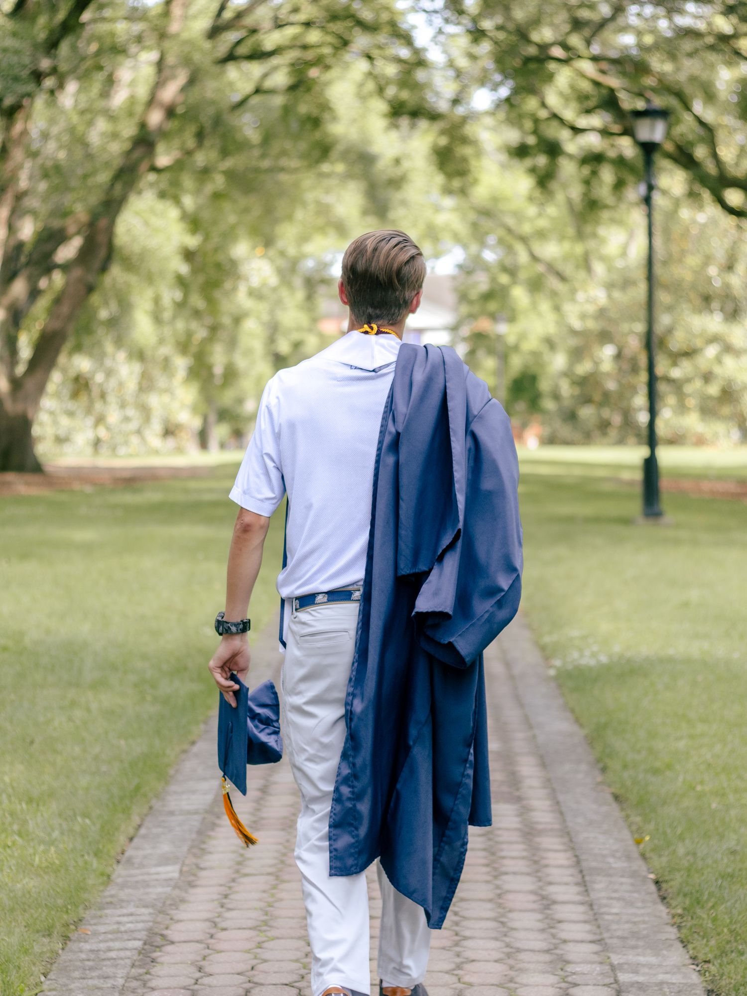 Graduate in a white polo and khakis walks a brick path under oak trees, carrying a navy gown and cap with a gold tassel.