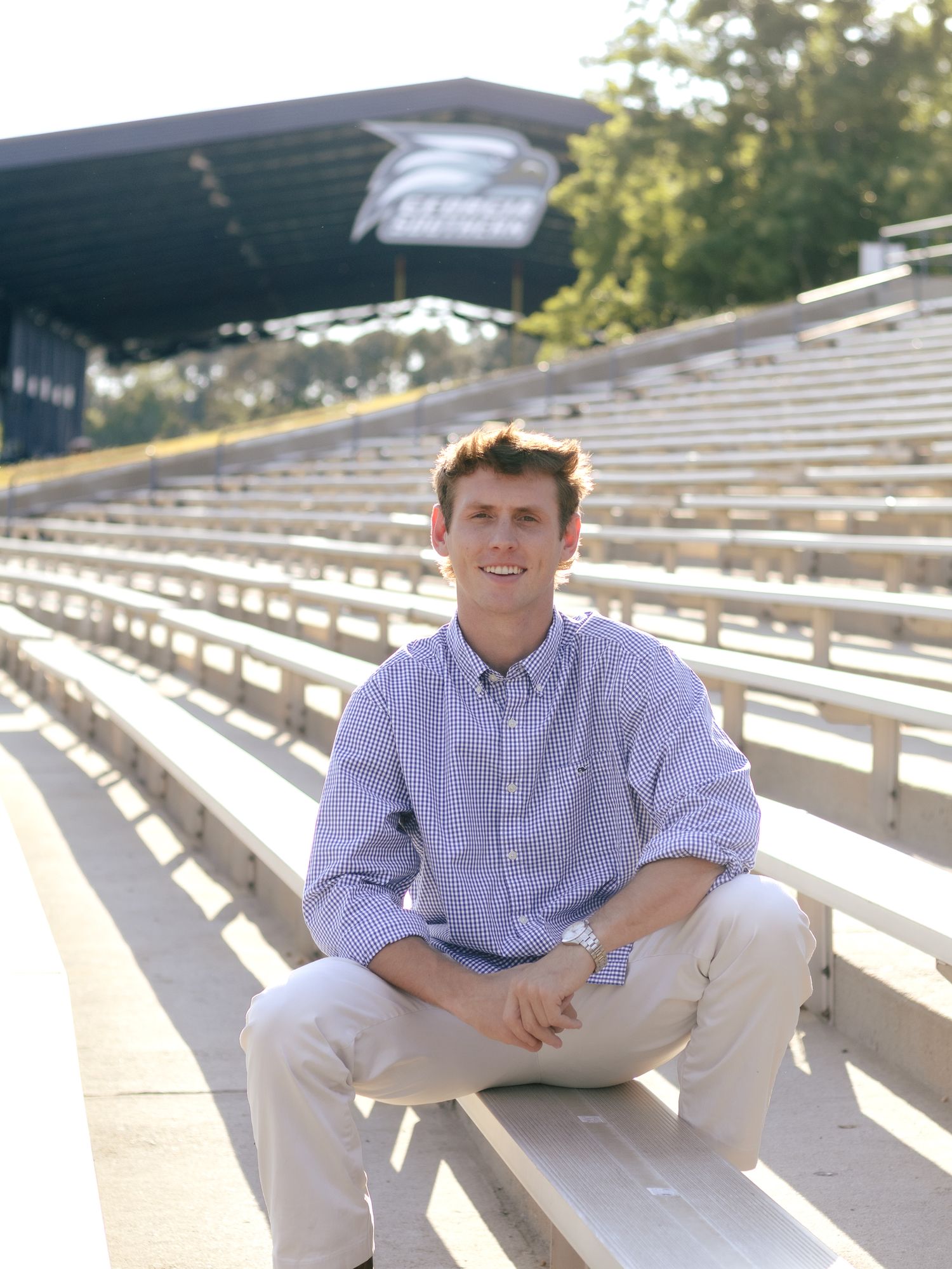 Smiling young man in a light blue button-up shirt and khaki pants sits on empty aluminum stadium bleachers on a sunny day.