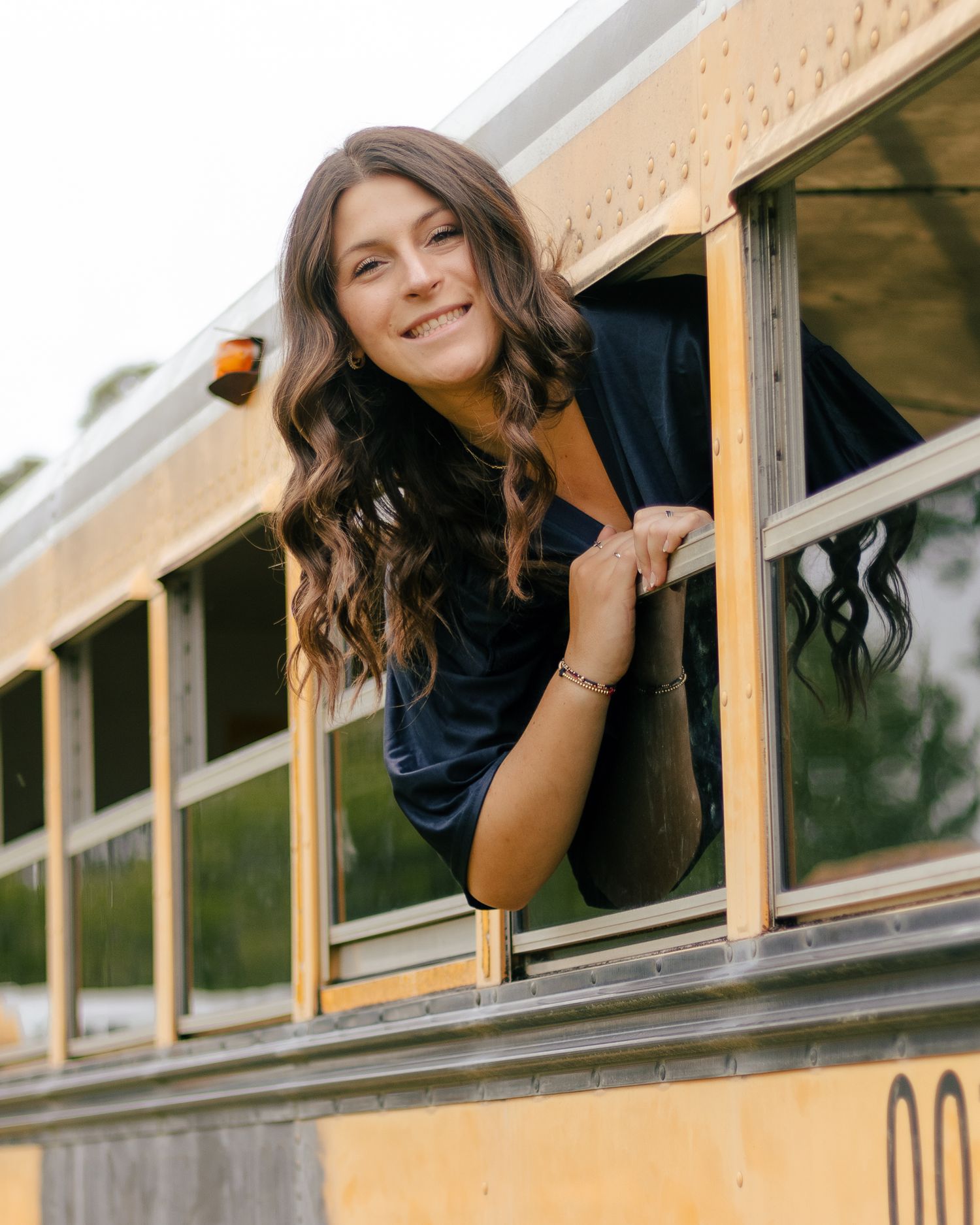 Smiling young woman with long wavy brown hair leans out of a yellow school bus window, resting her arms on the frame outdoors.