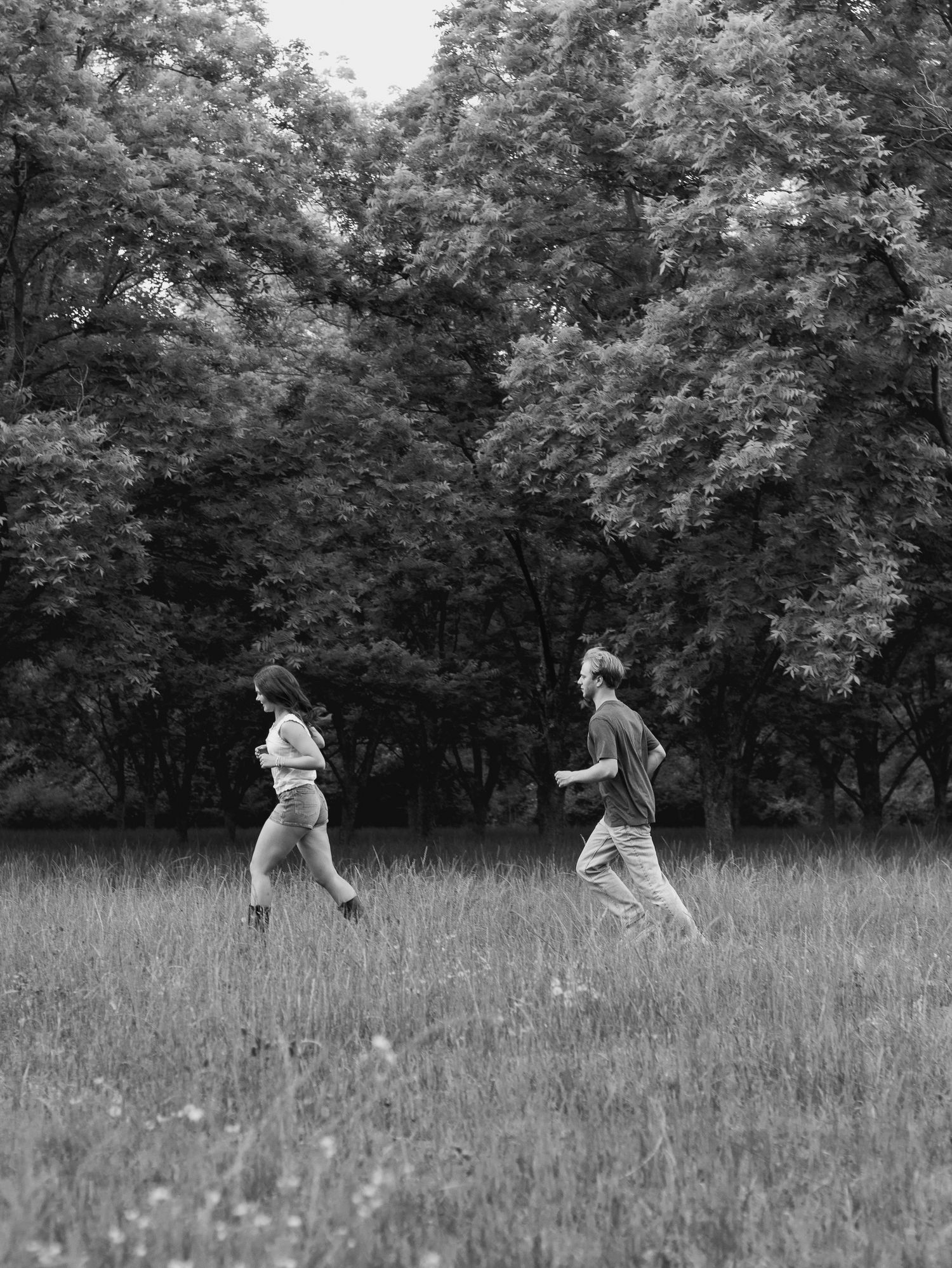 Black and white photo of two people running side by side through a tall grass meadow with dense leafy trees in the background.