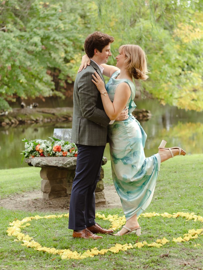 A couple embraces beside a pond as the woman kicks up her heel, standing in a circle of yellow flower petals near a floral stone bench.