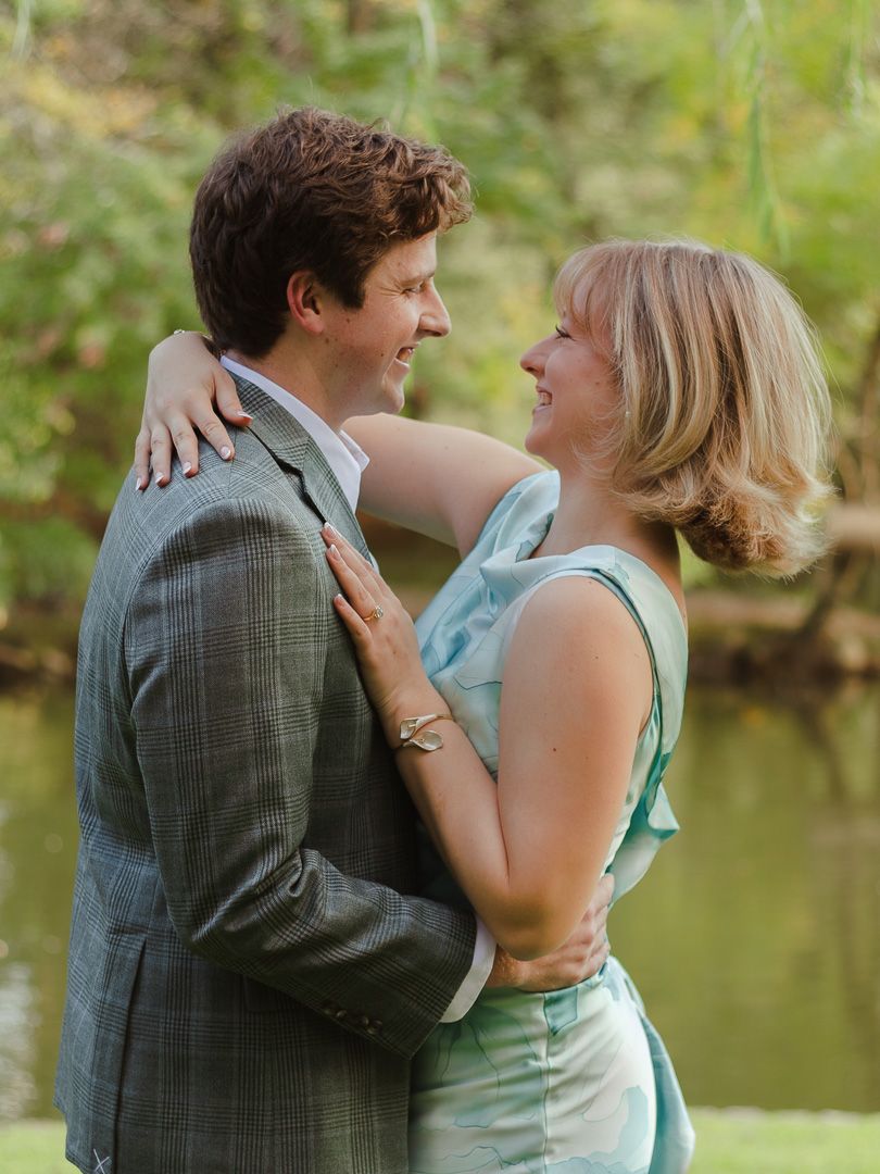 A smiling couple embraces by a tranquil pond, the man in a gray plaid blazer and the woman in a mint green dress.