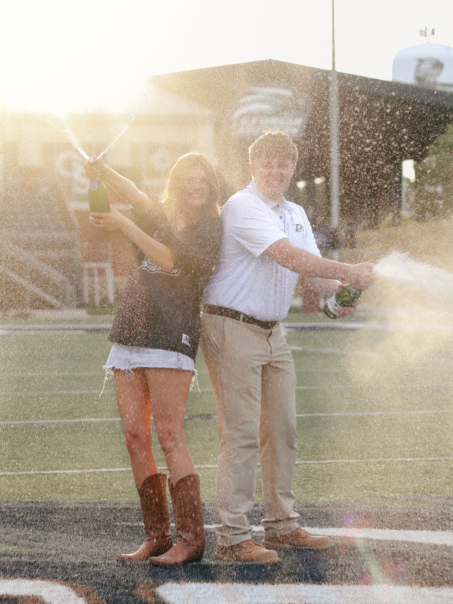 Young couple spraying champagne on a football field at sunset, she in cowboy boots and he in a white polo and khakis.