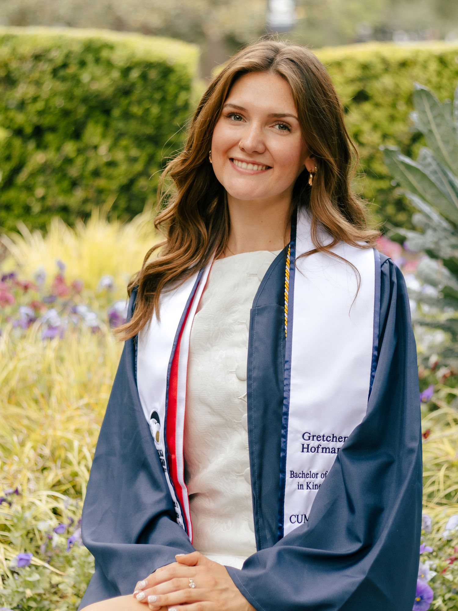 Smiling graduate in a navy gown and white stole poses outdoors in front of a colorful garden with green hedges and purple flowers.