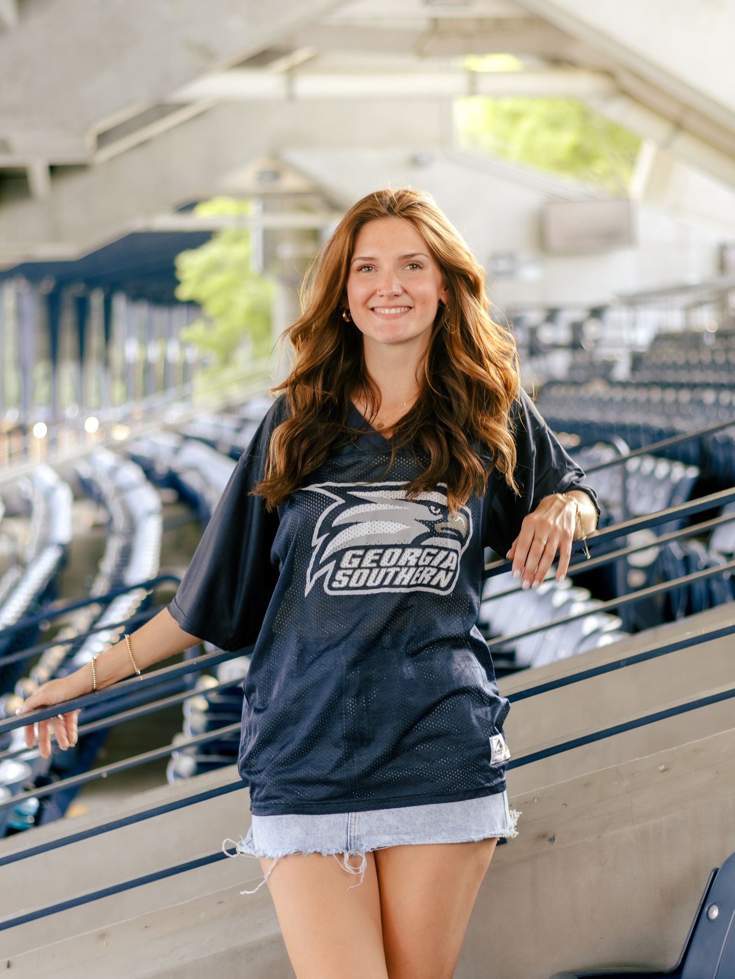 Smiling young woman with long wavy hair wears a navy Georgia Southern jersey and denim shorts while posing in an empty stadium.