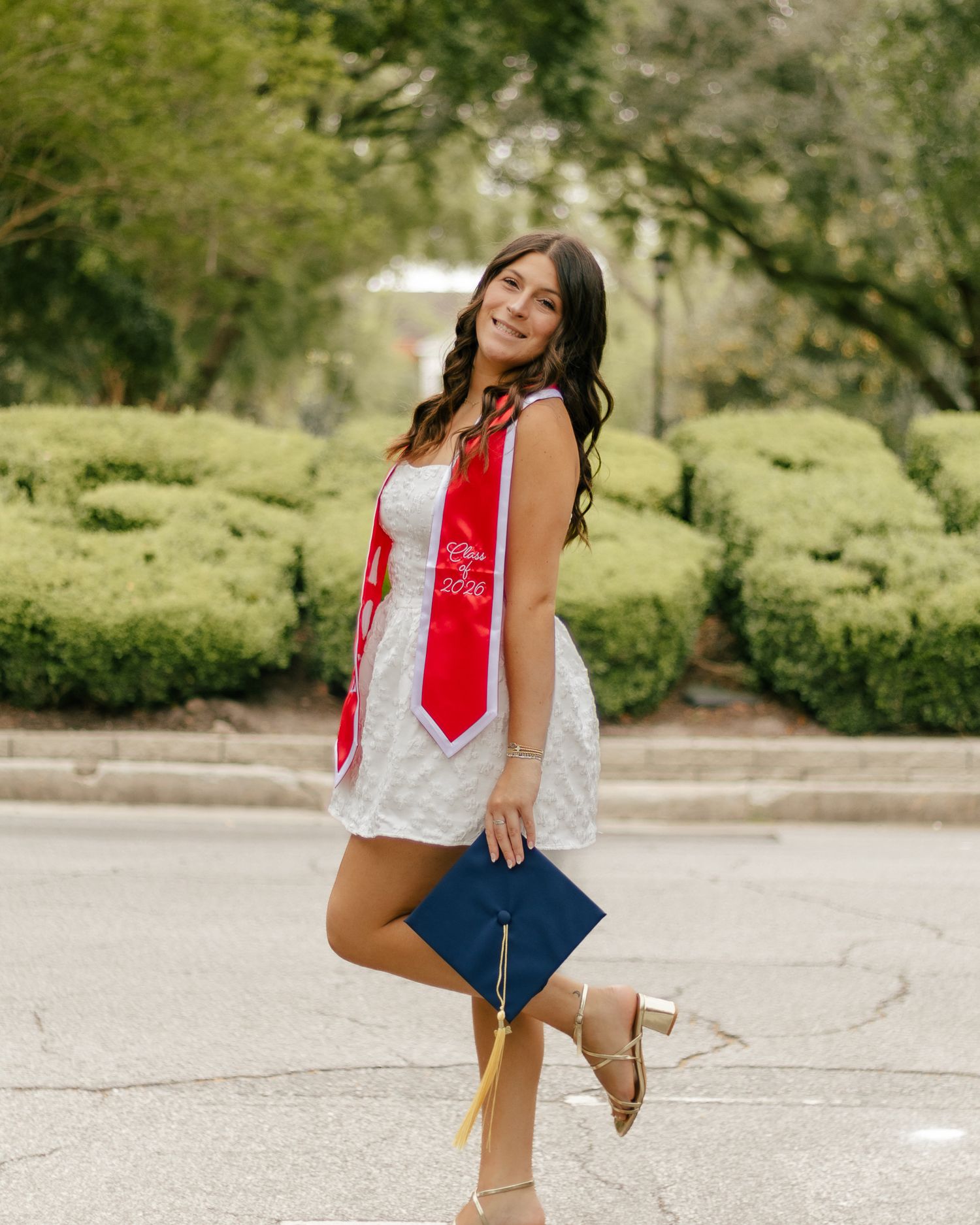 Smiling graduate in a white lace dress and red graduation stole holds a navy cap while posing on a tree-lined street.