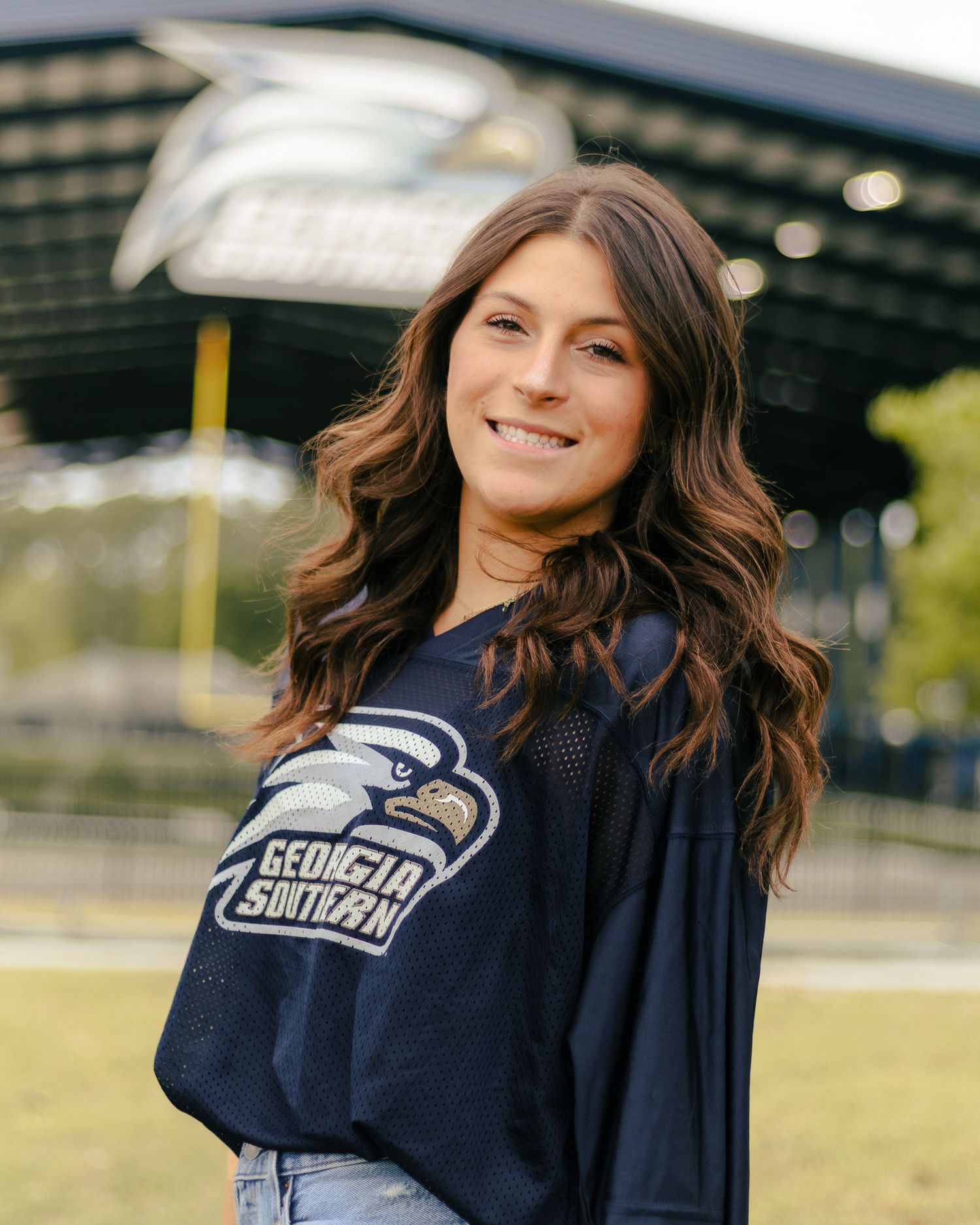 Smiling woman with long wavy brown hair wears a navy Georgia Southern eagle jersey while posing outdoors in front of a stadium.