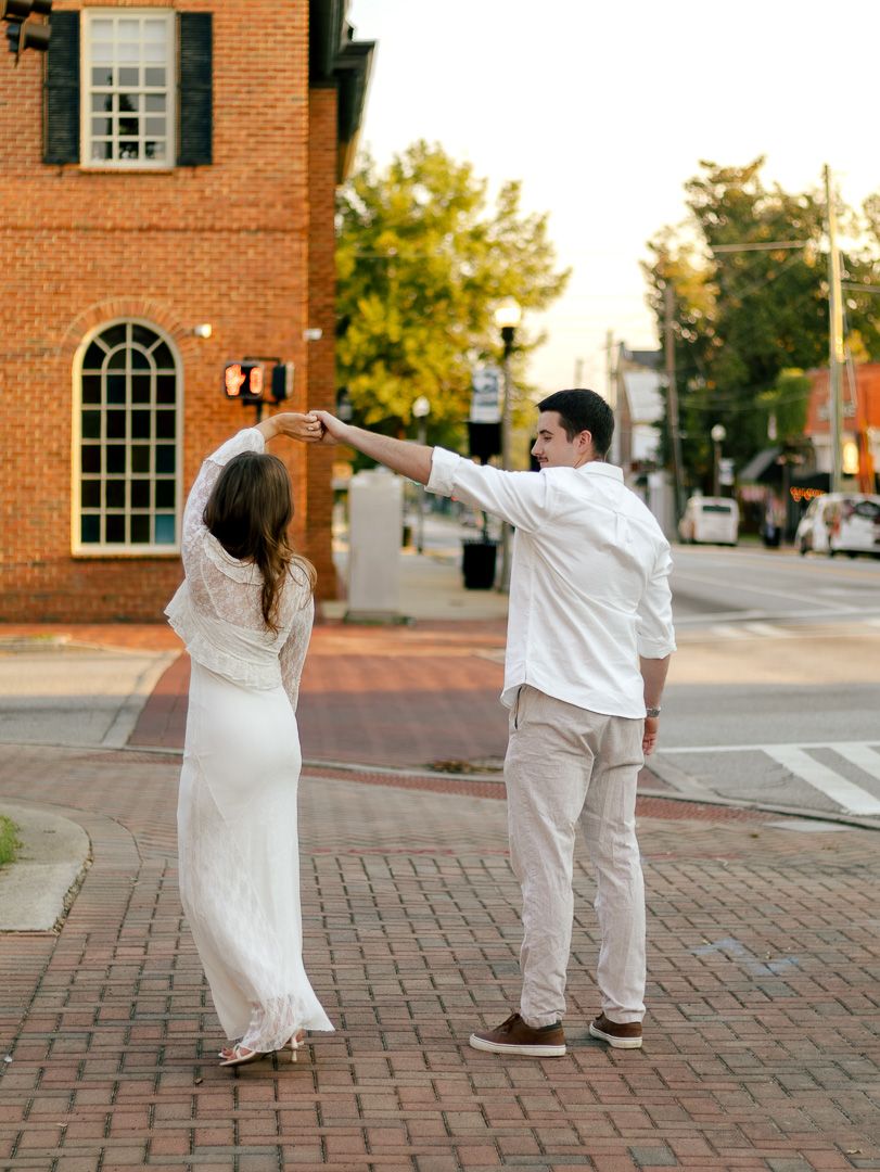 Couple in white outfits dancing on a brick-paved street corner beside a historic red brick building at sunset.