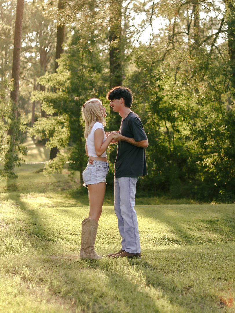 Young couple holding hands and facing each other on a sunlit grassy lawn surrounded by tall trees, wearing denim and cowboy boots.