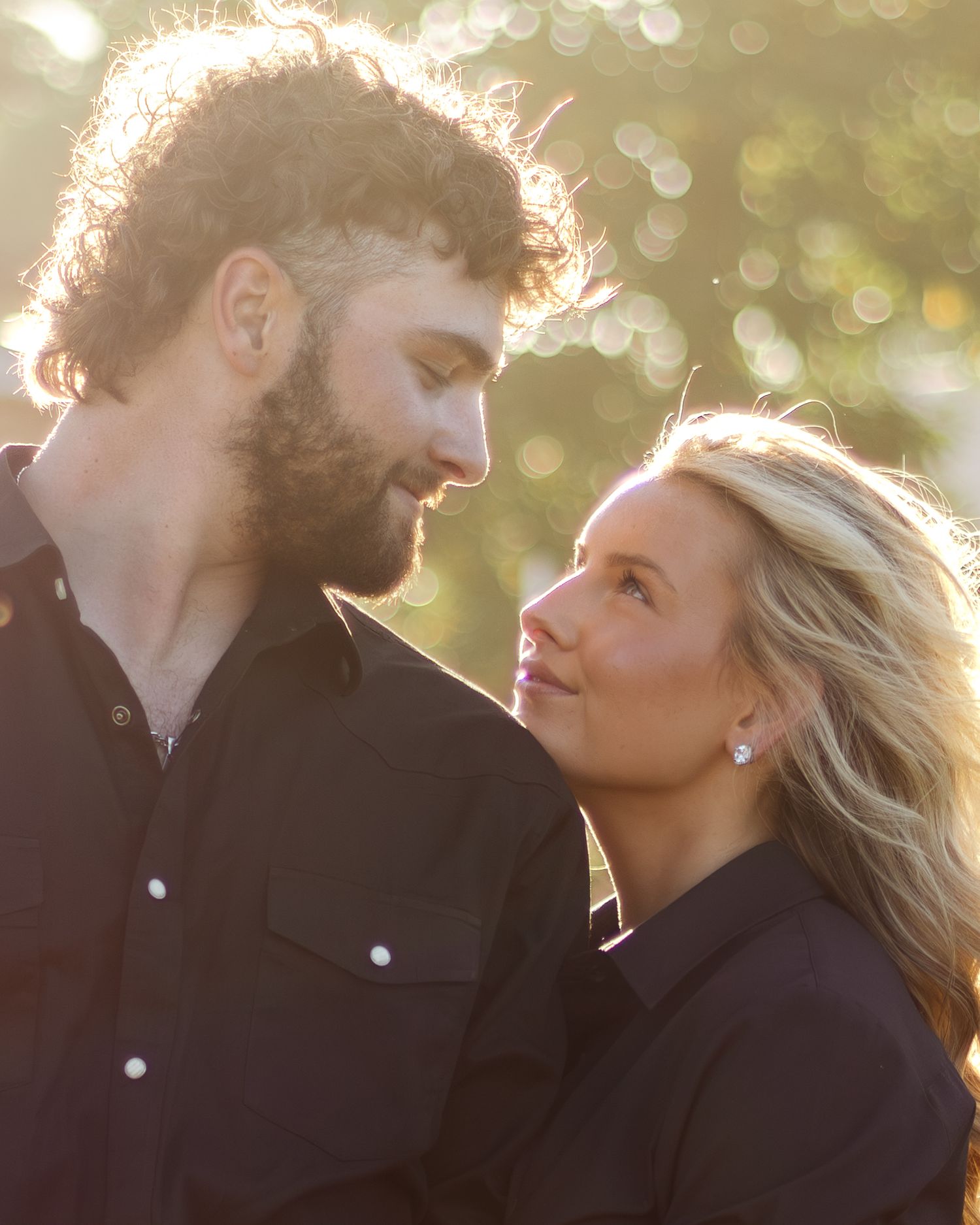 Bearded man and blonde woman in dark button-up shirts gaze lovingly at each other in warm golden sunlight with a blurred green background.