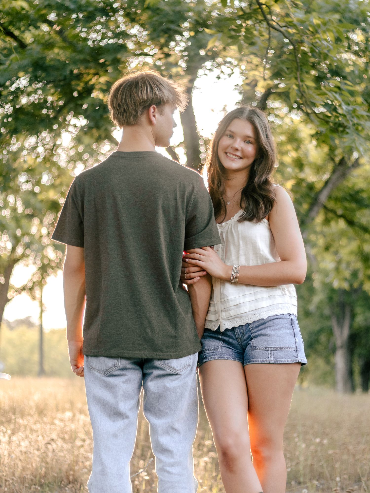 Two teenagers stand close together in a sunlit grassy field with trees behind them, the girl smiling in a white tank top and denim shorts.