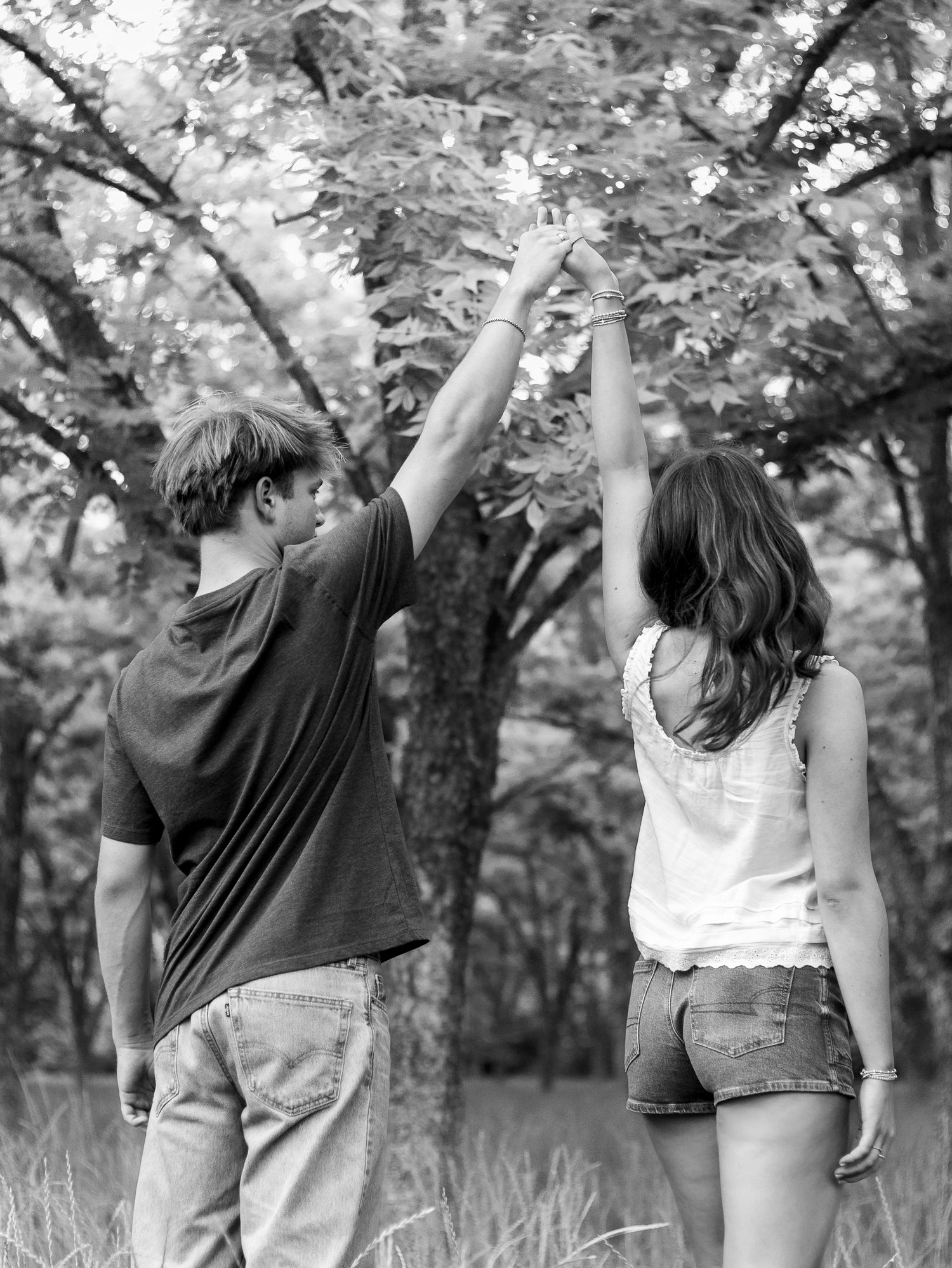 Black and white photo of a couple from behind holding raised hands while standing together in a grassy forest clearing.