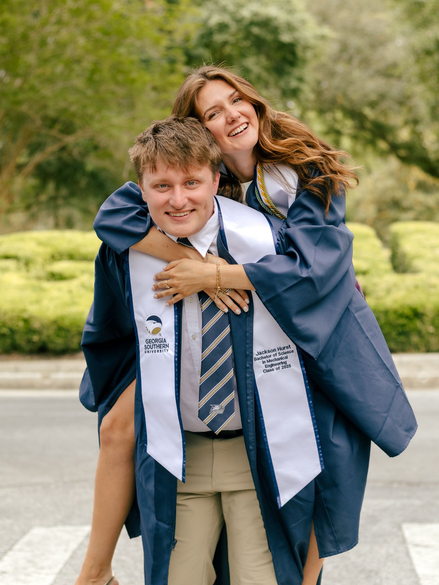 Smiling graduate in a navy gown and white Georgia Southern stole gets a piggyback hug from a woman in graduation attire outdoors.