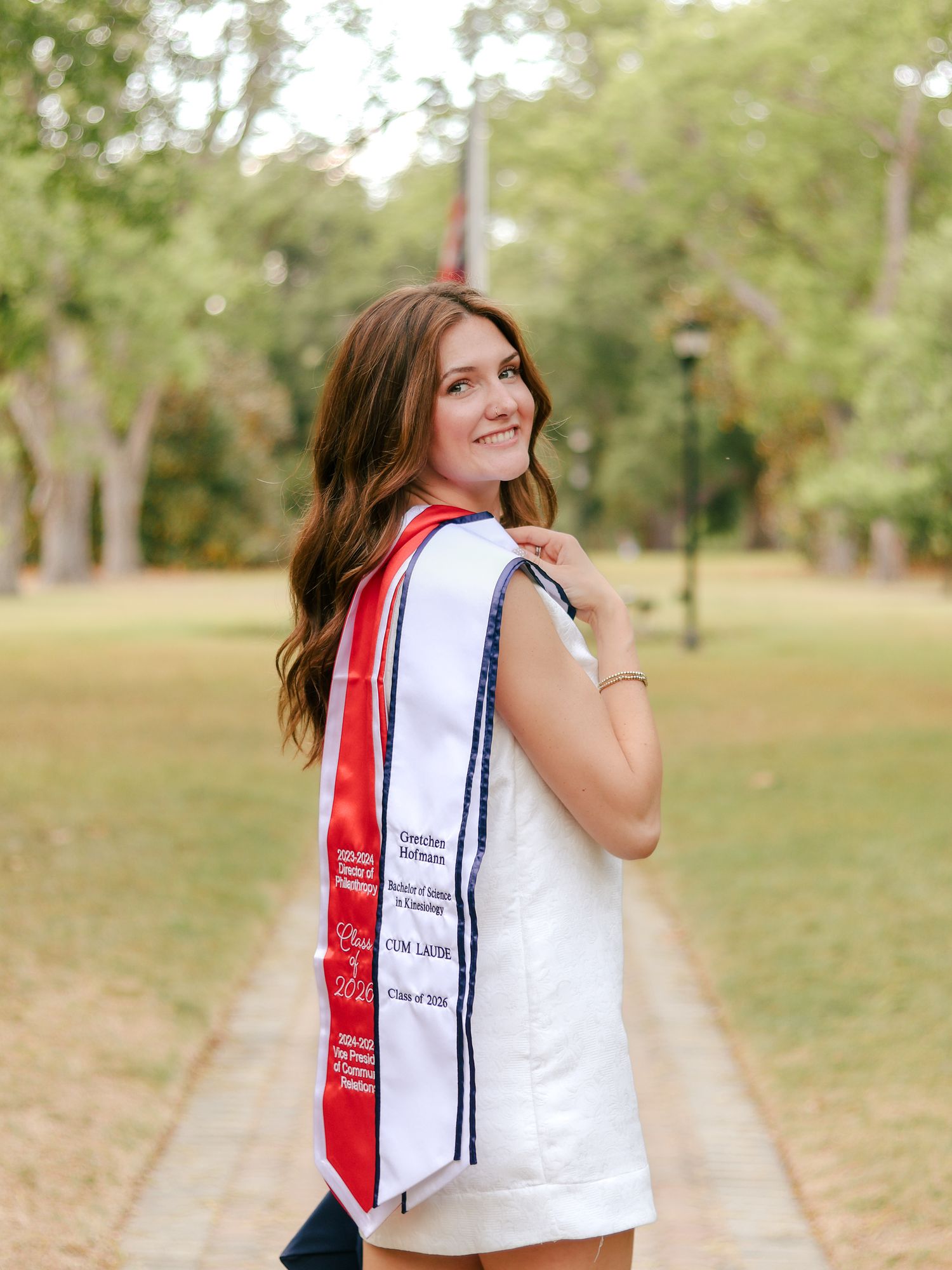 Smiling graduate with long brown hair wears a white dress and red, white and navy graduation stole while posing on a tree-lined brick path.