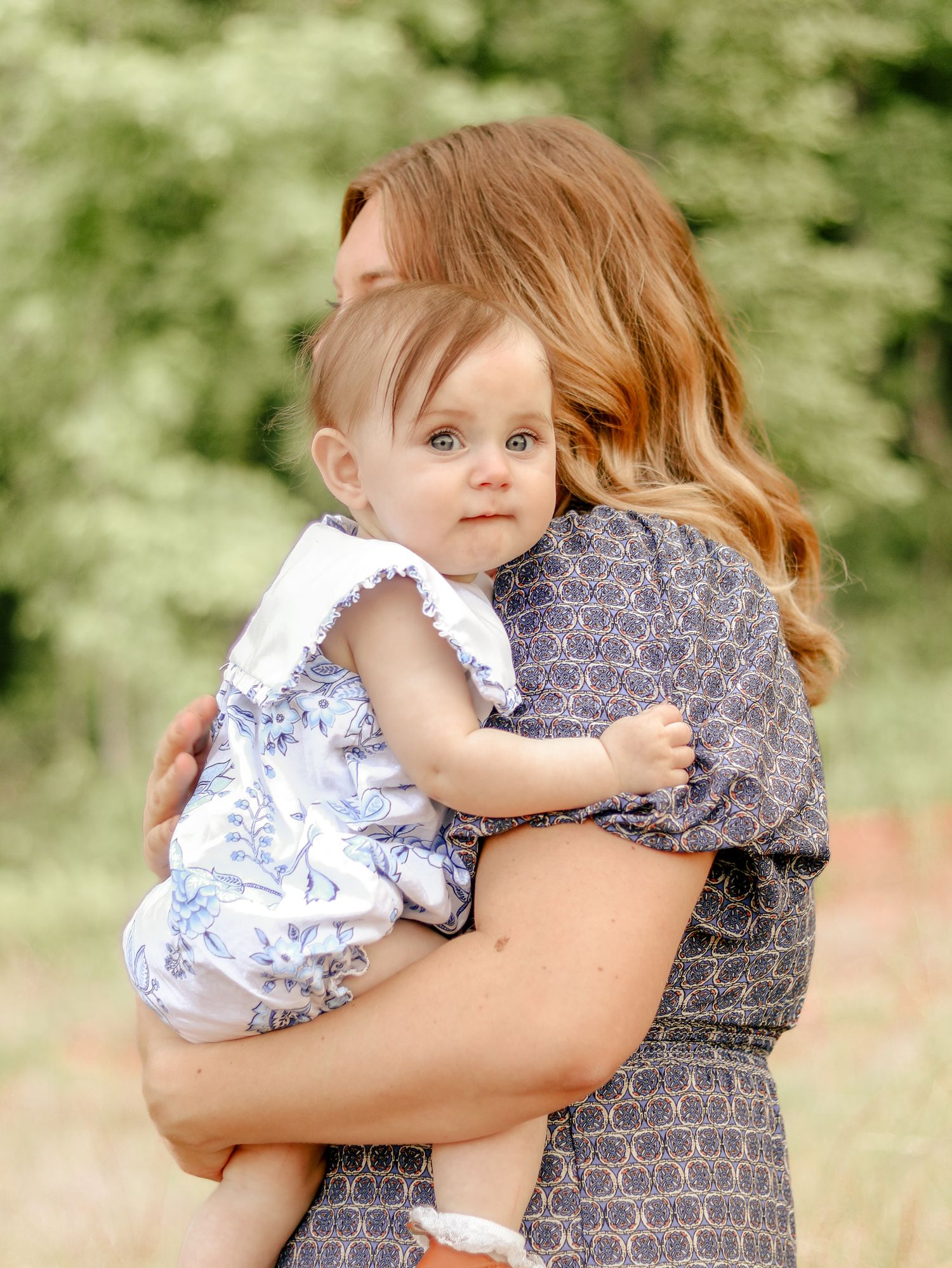 A woman with auburn hair holds a baby in a blue floral romper outdoors, with soft green trees in the background.