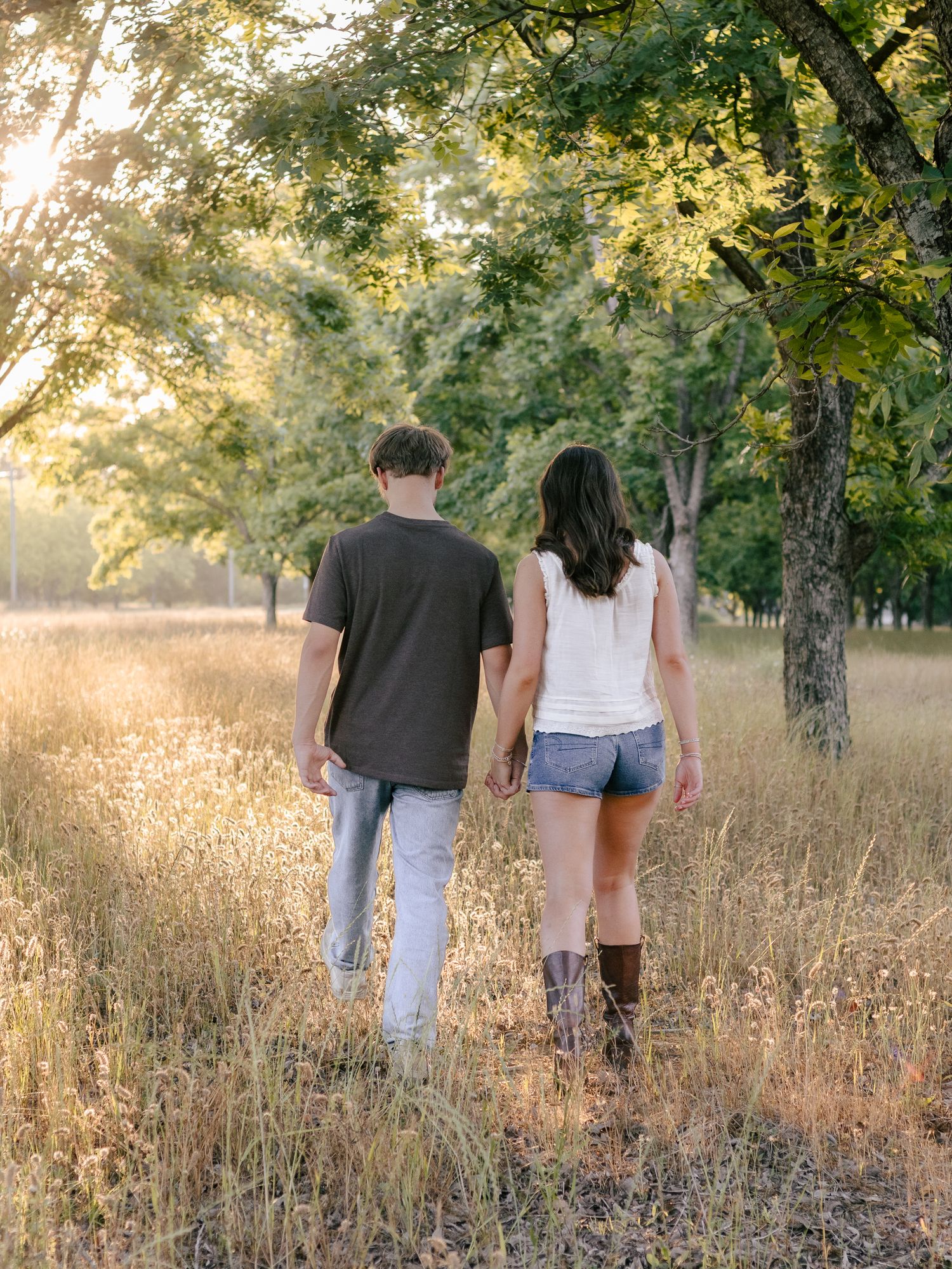 Couple holding hands walking away through a golden grassy field under sunlit trees in warm evening light.