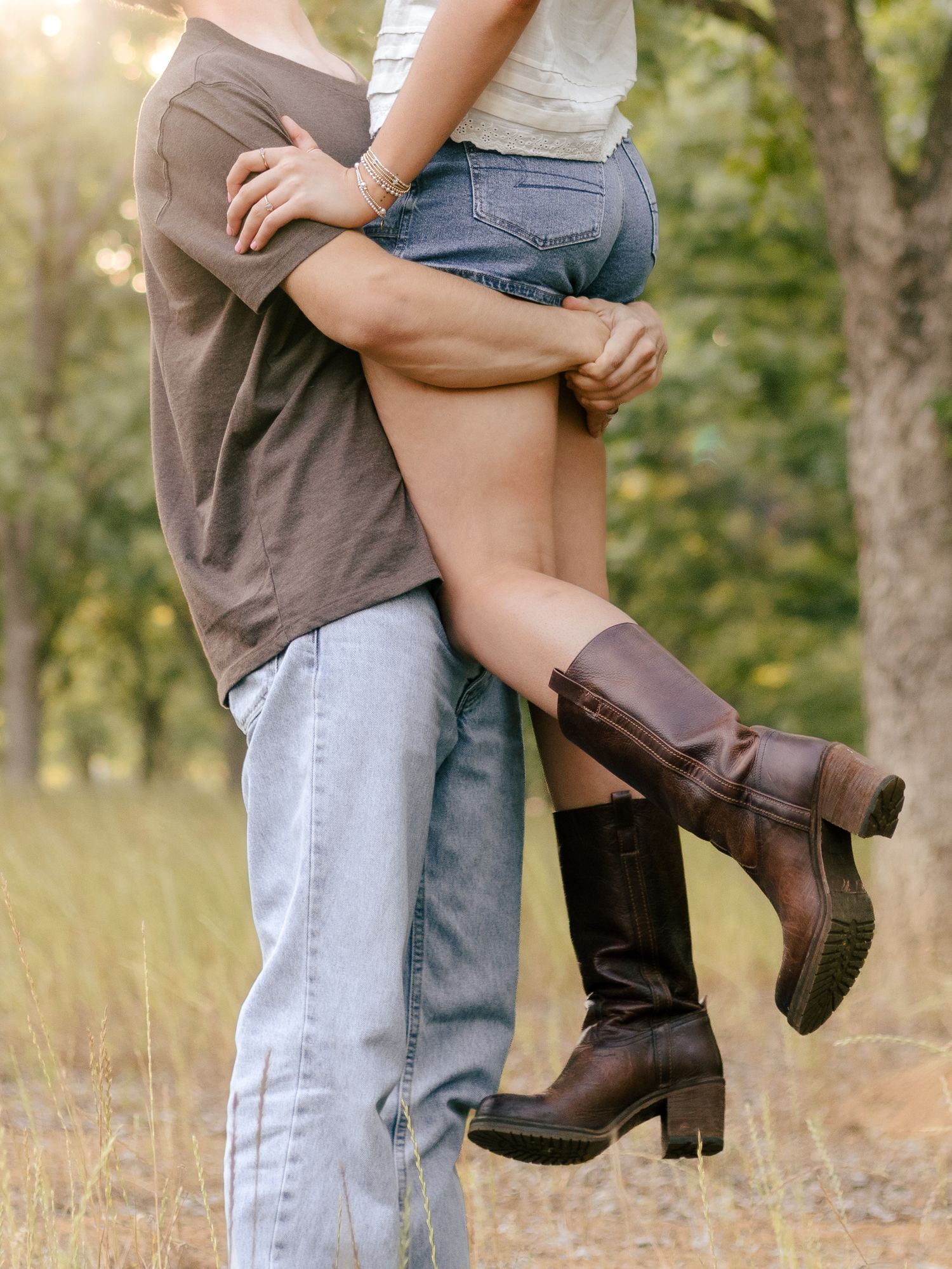 Man in a brown t-shirt and jeans lifts a woman in denim shorts and brown cowboy boots in a sunny grassy field with trees.