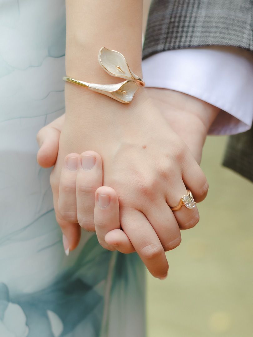 Close-up of a couple holding hands, showing a diamond ring and an elegant gold calla lily bracelet on the woman's wrist.
