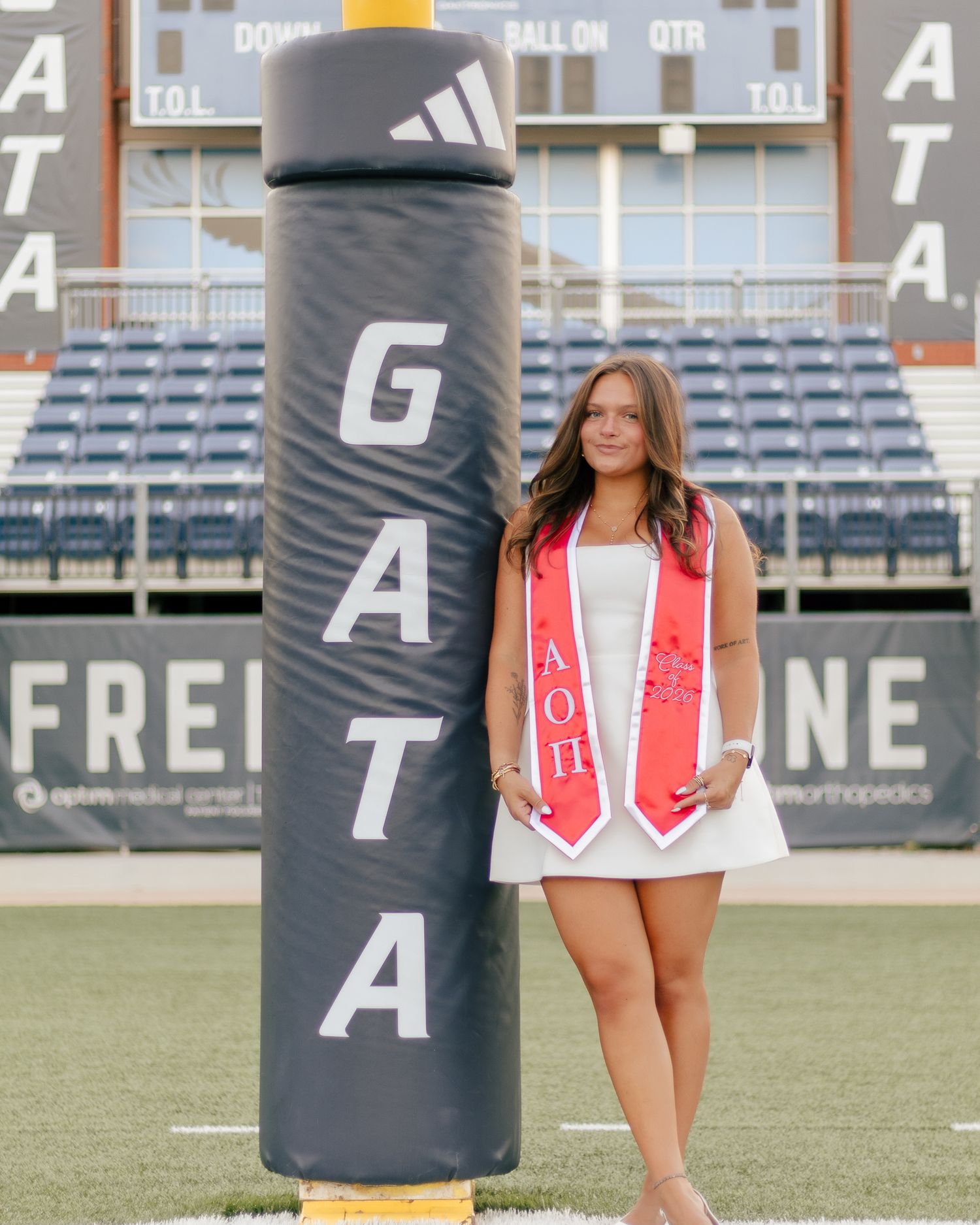 Smiling graduate in a white dress and red Alpha Omicron Pi stole poses beside a padded goalpost at a football stadium.