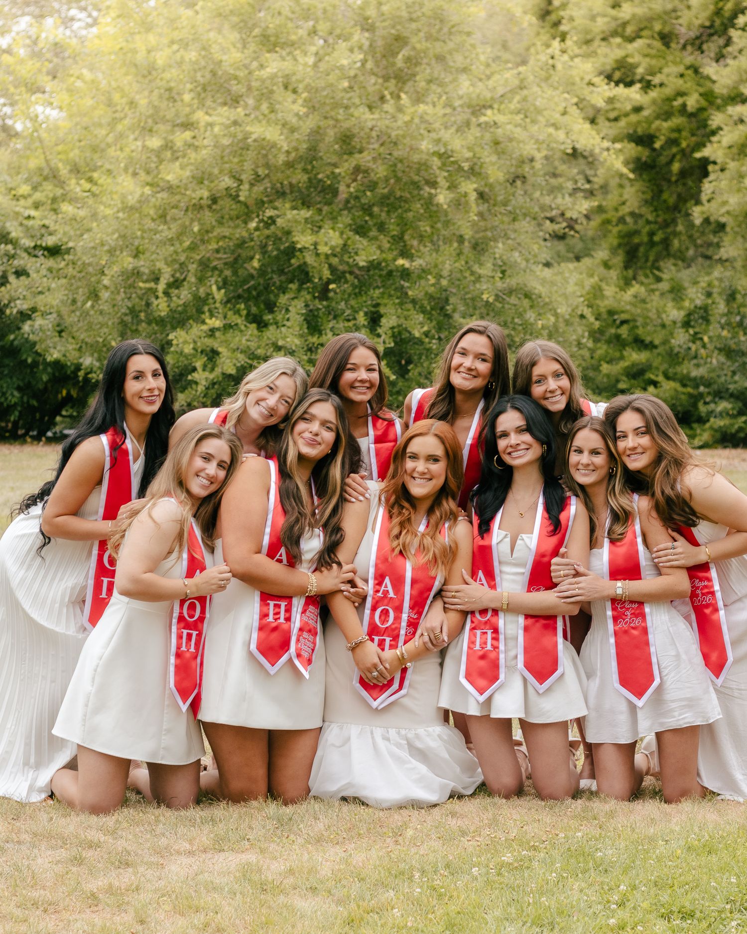 Group of smiling women in white dresses and red sashes pose together on a grassy lawn with green trees in the background.