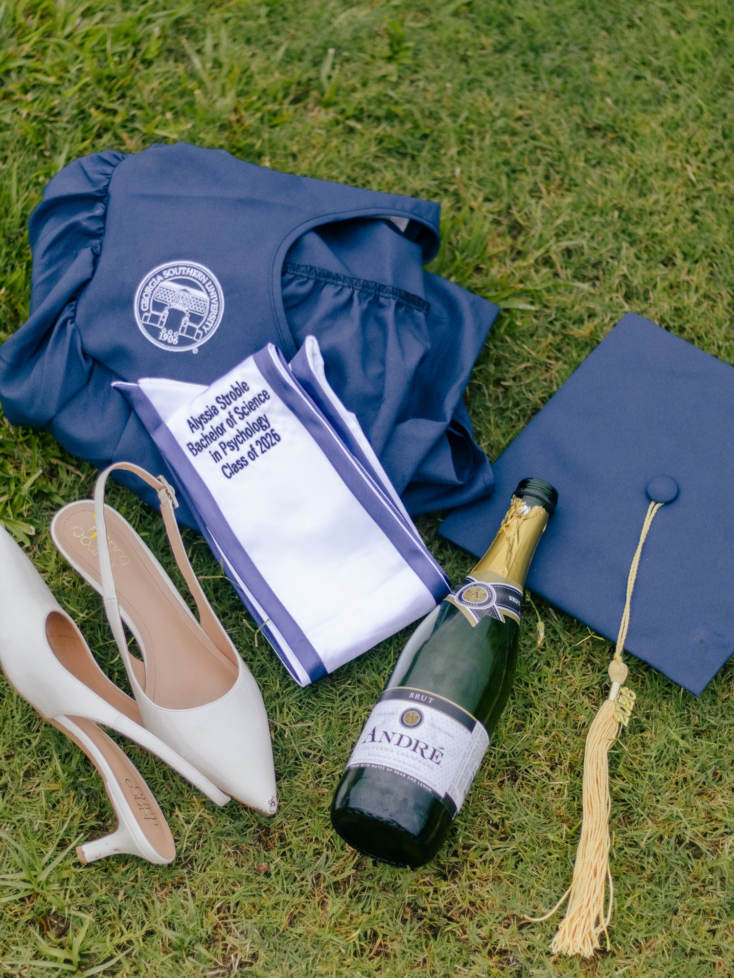 Navy graduation gown, sash, cap with gold tassel, white heels, and a bottle of Andre champagne arranged on green grass.