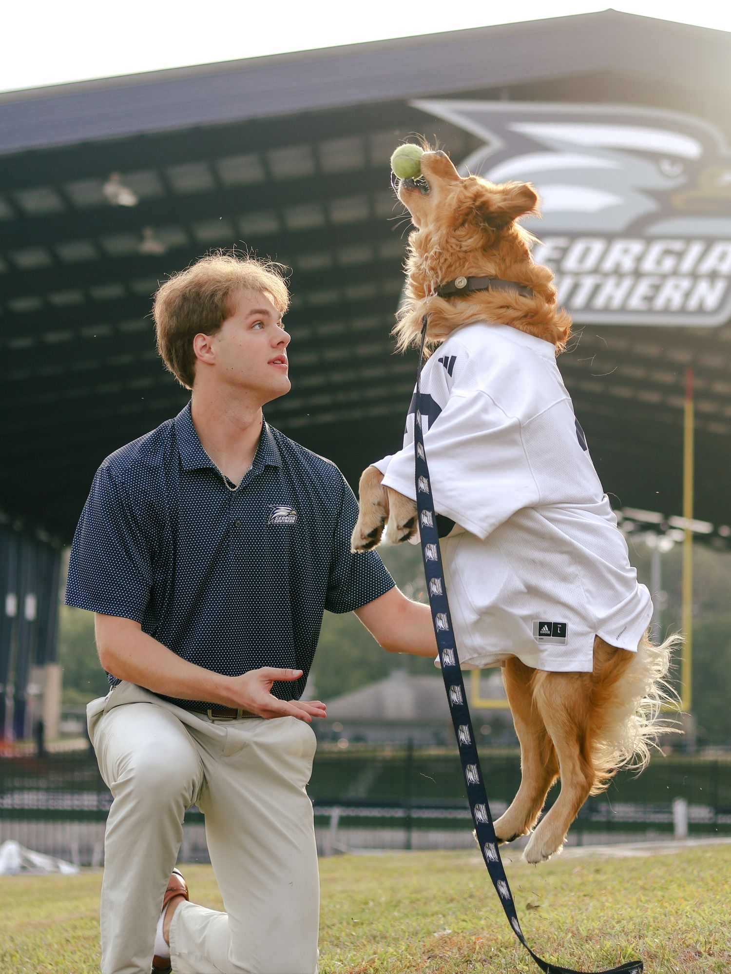 A man kneels on the grass as a golden retriever in a white jersey leaps with a tennis ball in its mouth at a Georgia Southern stadium.