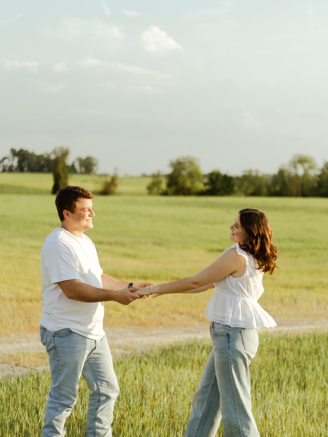 Smiling couple in white tops and jeans holding hands and dancing in a sunny green field with trees under a soft blue sky.