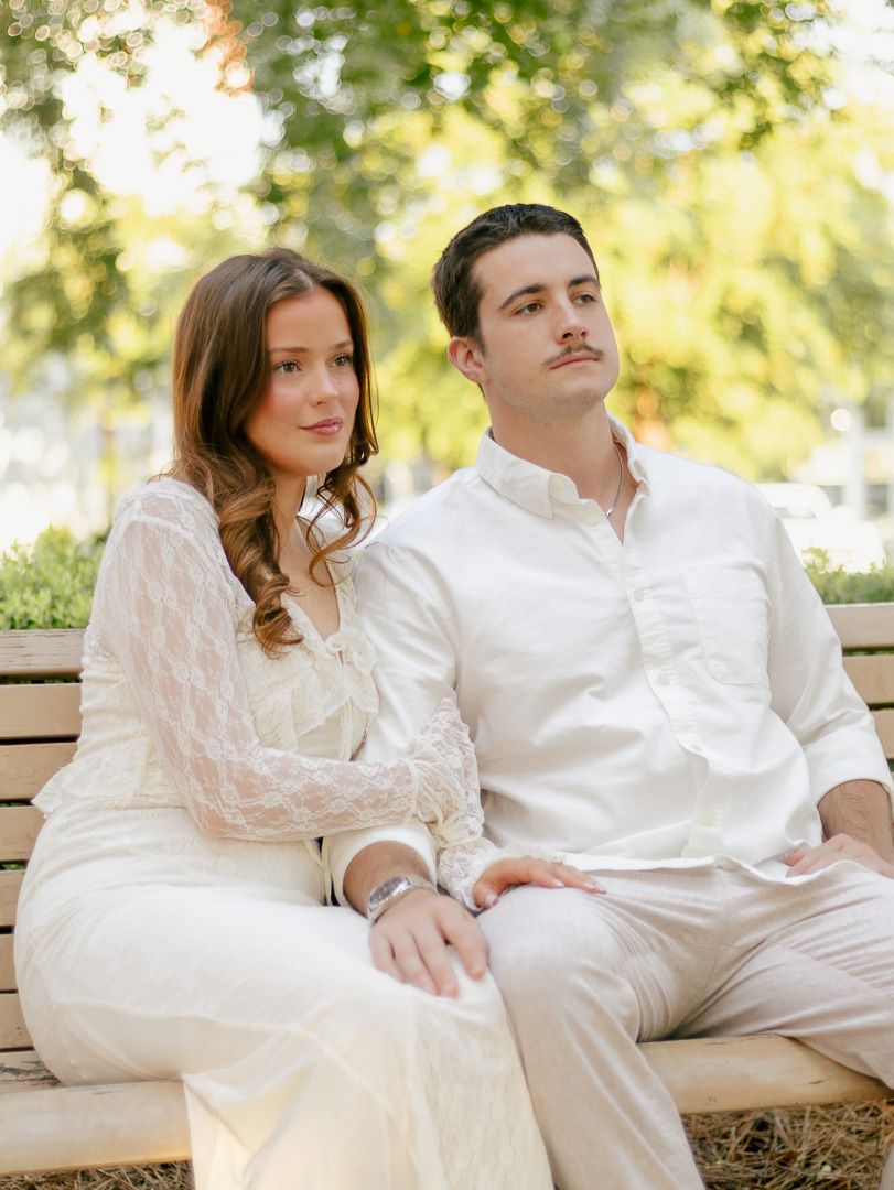 Couple in matching cream outfits sitting close together on a wooden park bench surrounded by green trees in soft natural light.