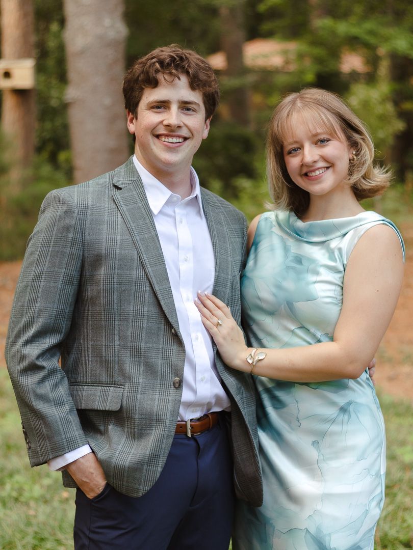 A smiling couple poses together outdoors among tall trees, the man in a gray plaid blazer and the woman in a blue dress with a bracelet.