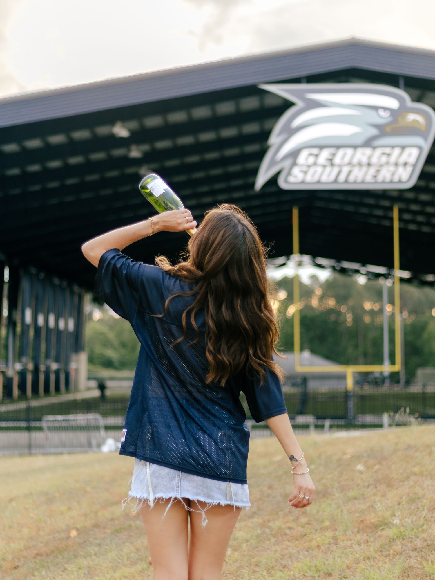Woman in a navy jersey and denim shorts tips a champagne bottle back near the Georgia Southern stadium sign.