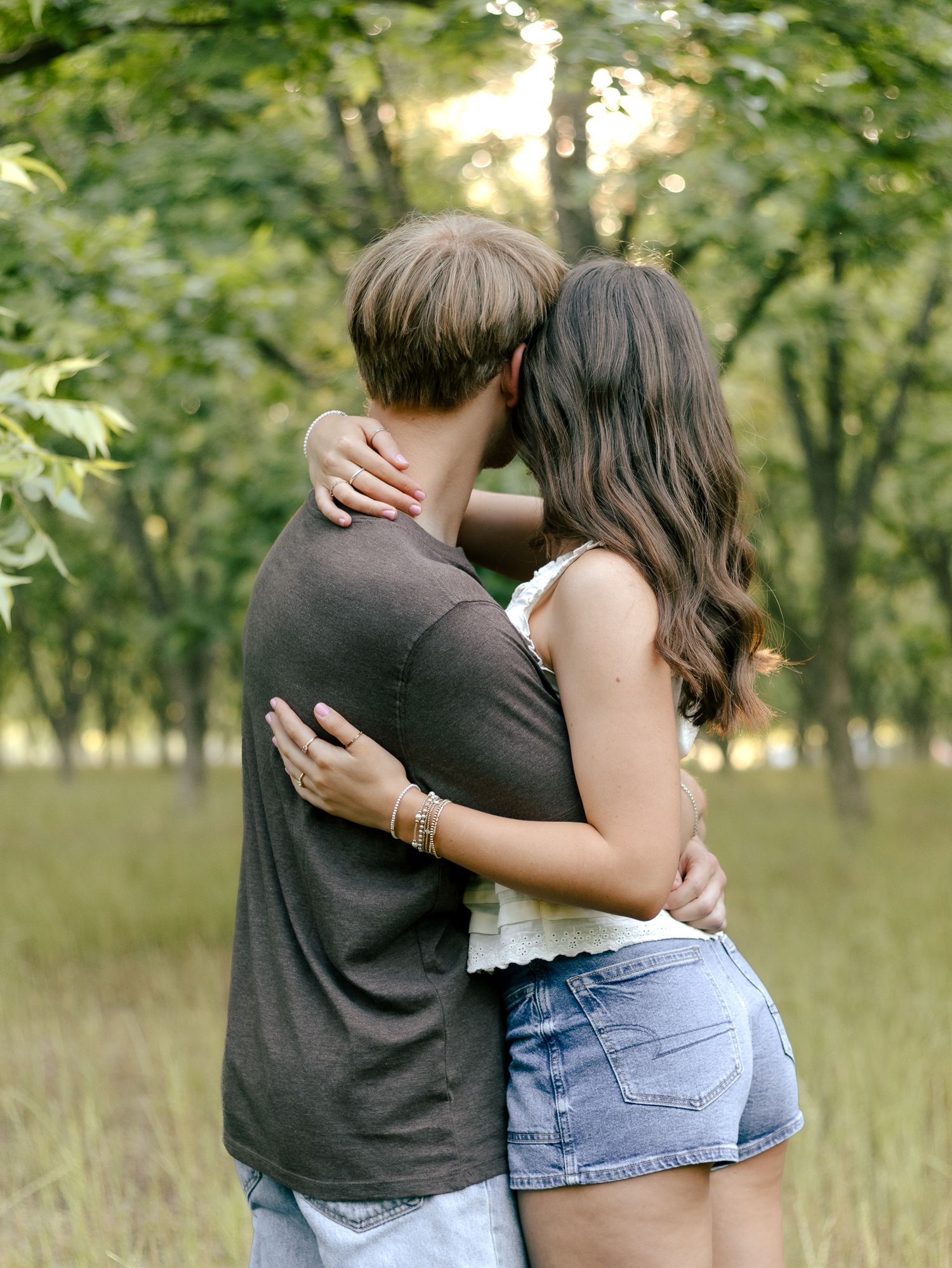 Couple embracing in a sunlit grassy field surrounded by trees, the woman in denim shorts resting her head on the man's shoulder.